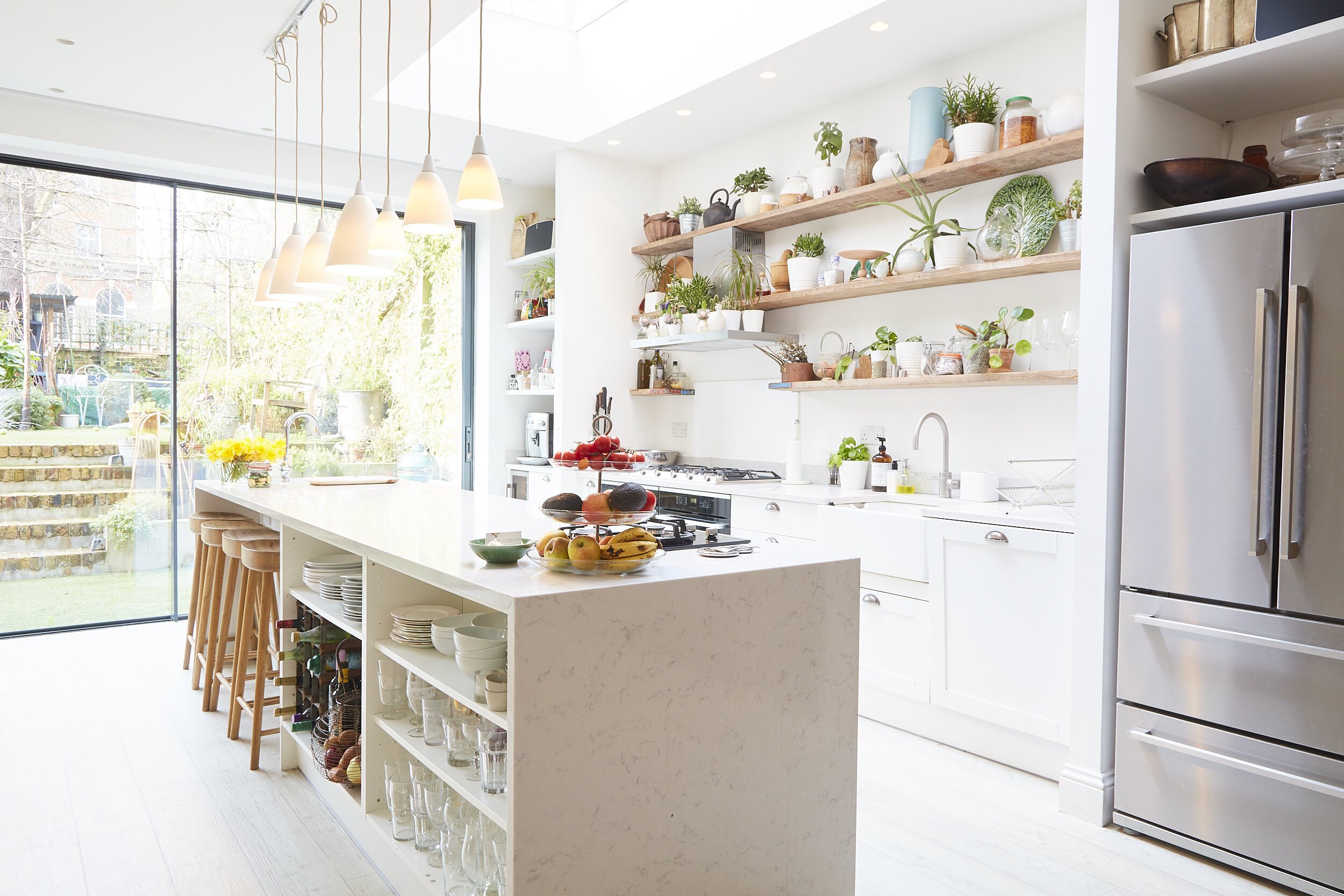 Bright, modern kitchen with white cabinets, open wooden shelves with plants and dishes, large sliding glass door leading to garden, and a kitchen island with stools and bowls of fruit.
