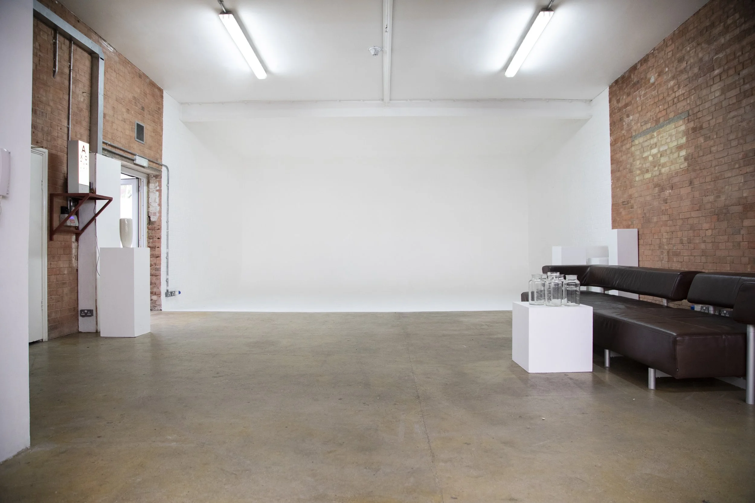 Empty art gallery with a brick wall on the right, a white wall in the background, and a black leather couch with glass jars on a white pedestal on the right side.