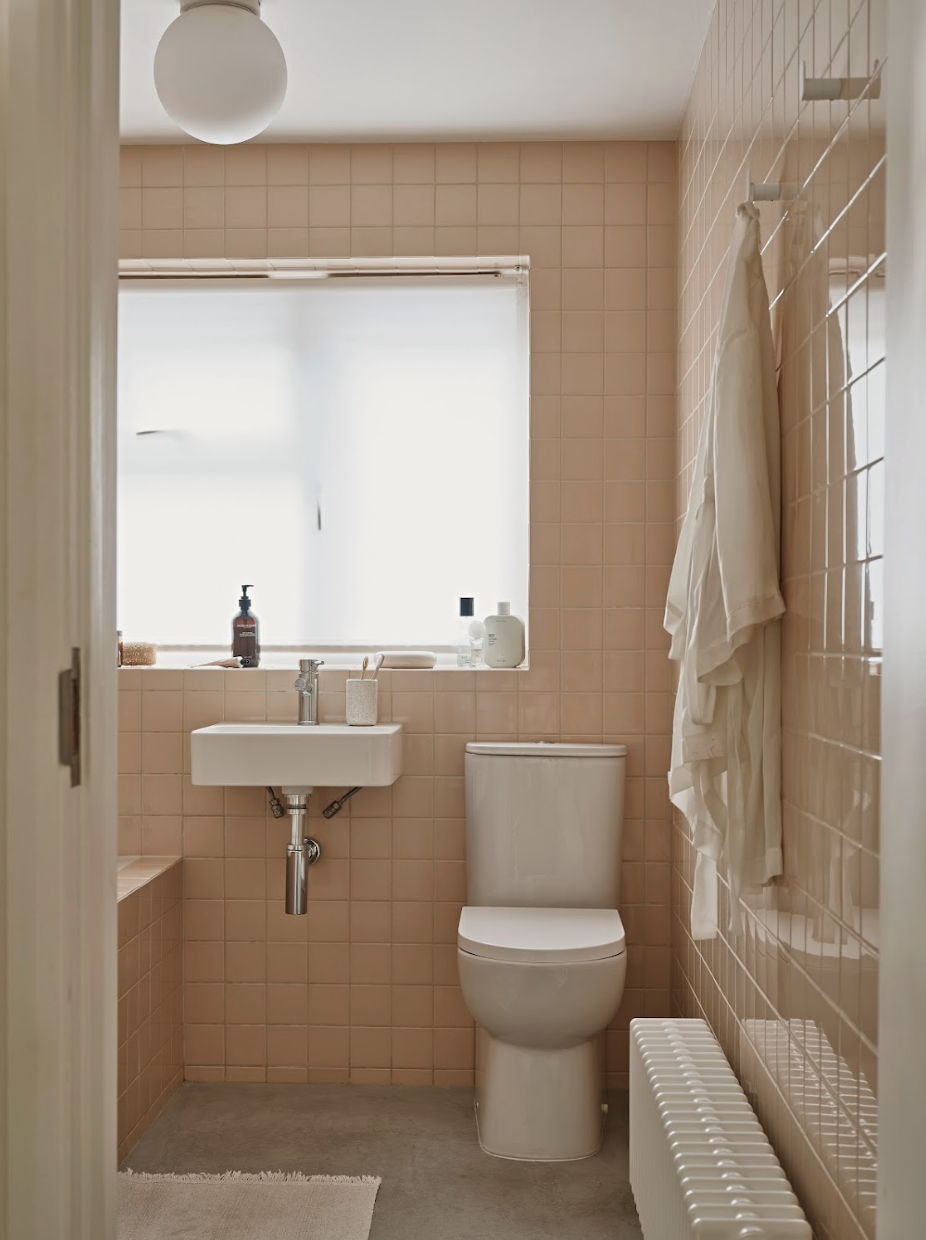 A small bathroom with peach-colored tiled walls, a window with frosted glass, a white toilet, a wall-mounted sink with two toothbrushes, and hanging white towels.
