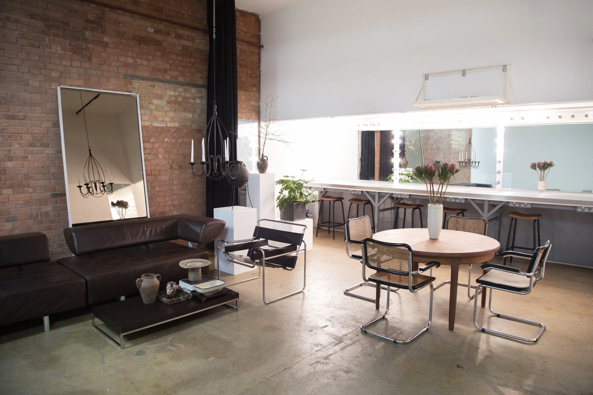 Living room with brick wall, mirror, black chandelier, brown leather sofa, chair, white pedestal with vase and branches, round table with flowers, bar counter with stools, large window.