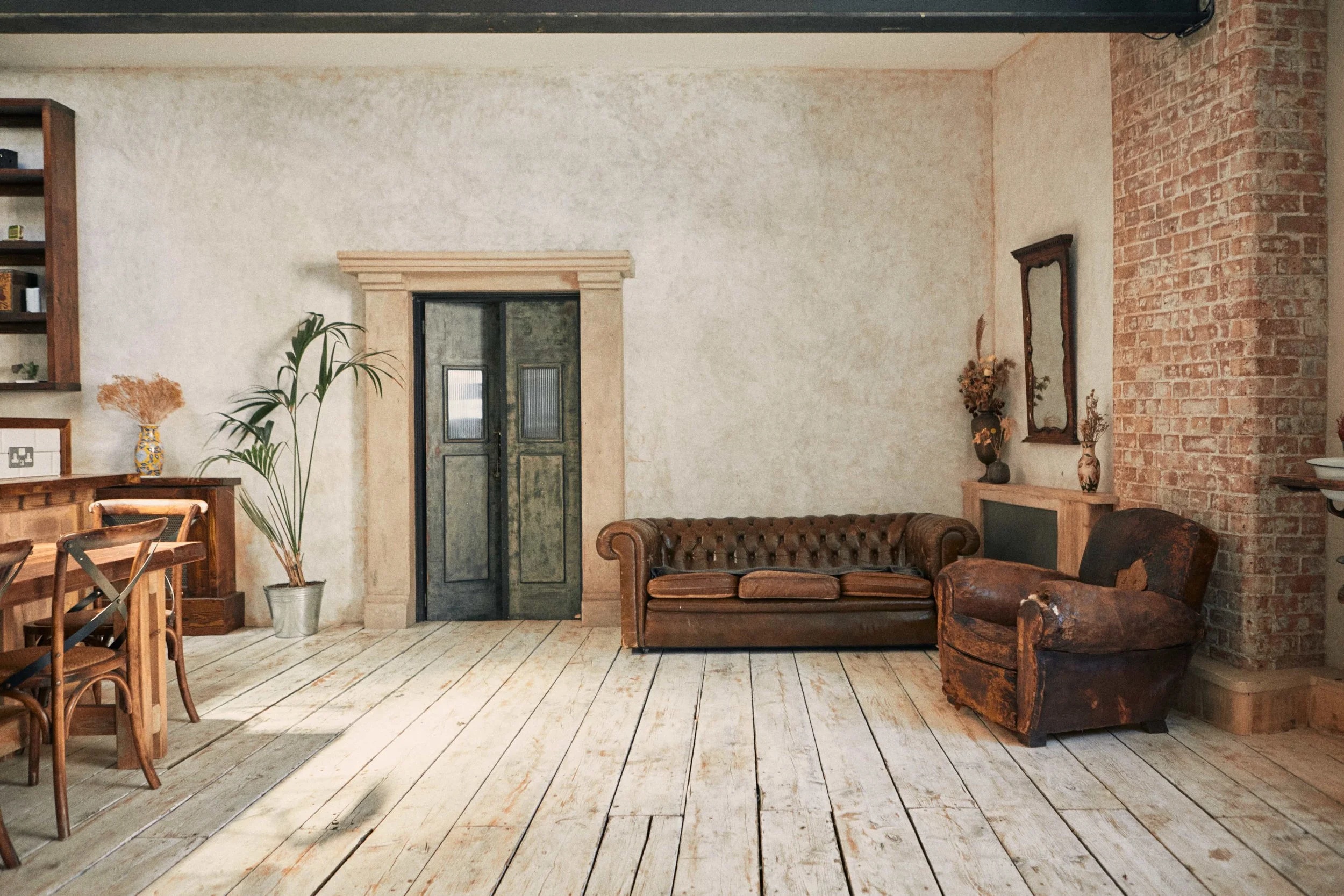 Living room with white wooden floor, brick and cream-colored walls, a dark green front door with a stone frame, a tall potted plant, a vintage leather sofa, a worn armchair, and decorative vases with dried flowers.