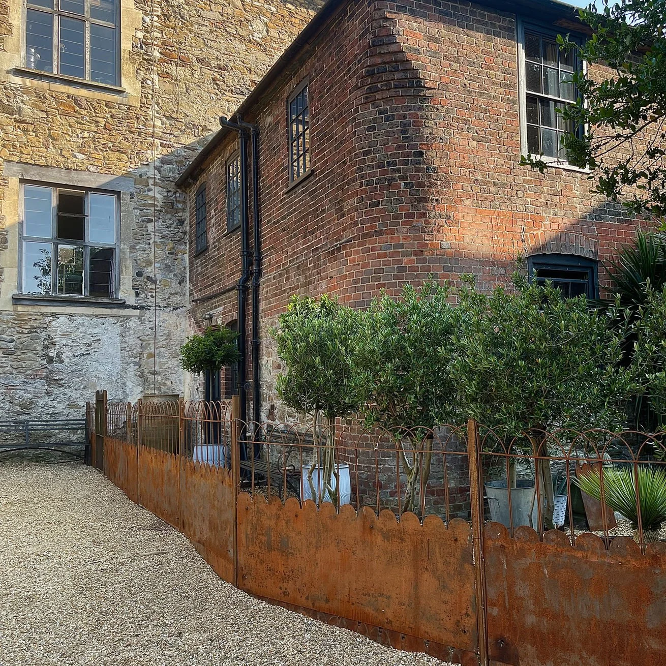 A rustic corner of an outdoor garden with green bushes, potted plants, and a rusted metal fence, next to brick and stone apartment building walls.