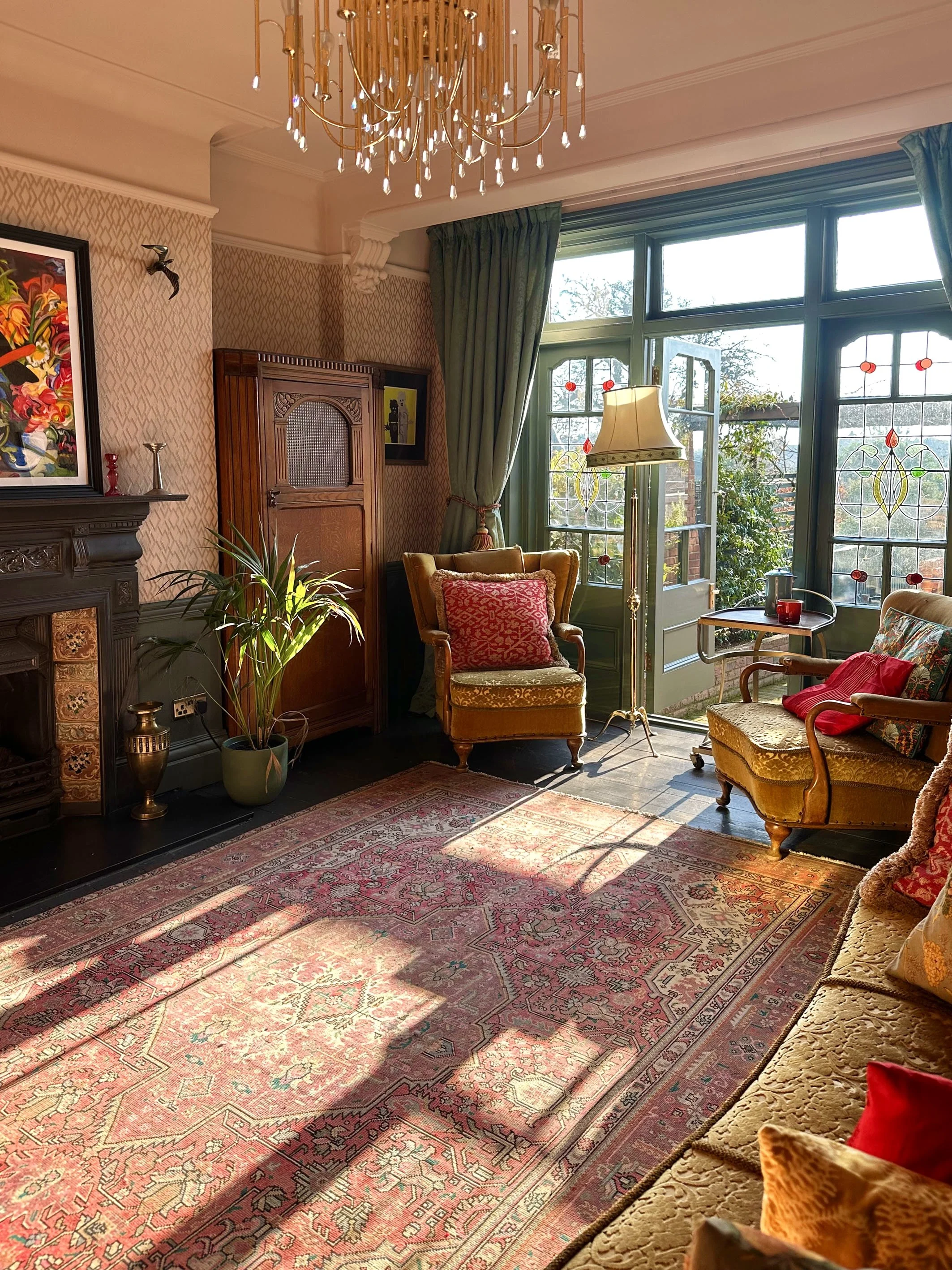 Sunlit living room with vintage yellow and patterned armchairs, a patterned oriental rug, potted plant, ornate wooden fireplace, stained glass windows, and a chandelier overhead.