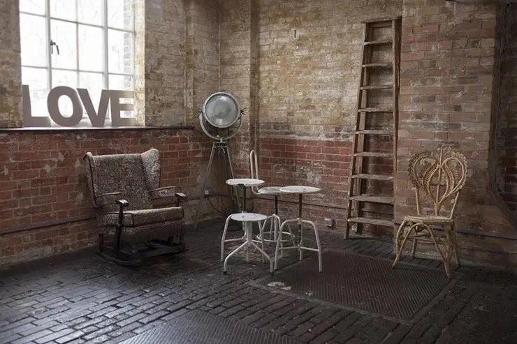 A cozy corner with vintage furniture including an armchair, two small white tables, a woven chair, and a ladder, set against brick walls with a window that has a 