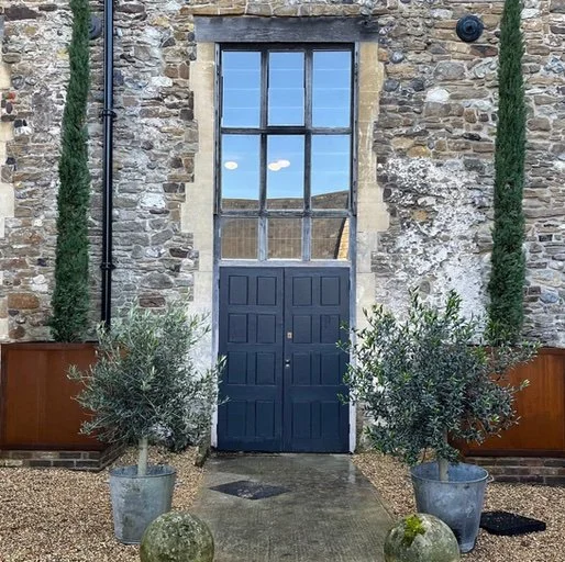 Stone building with large windowed door, flanked by two potted plants, and two tall narrow trees on either side.