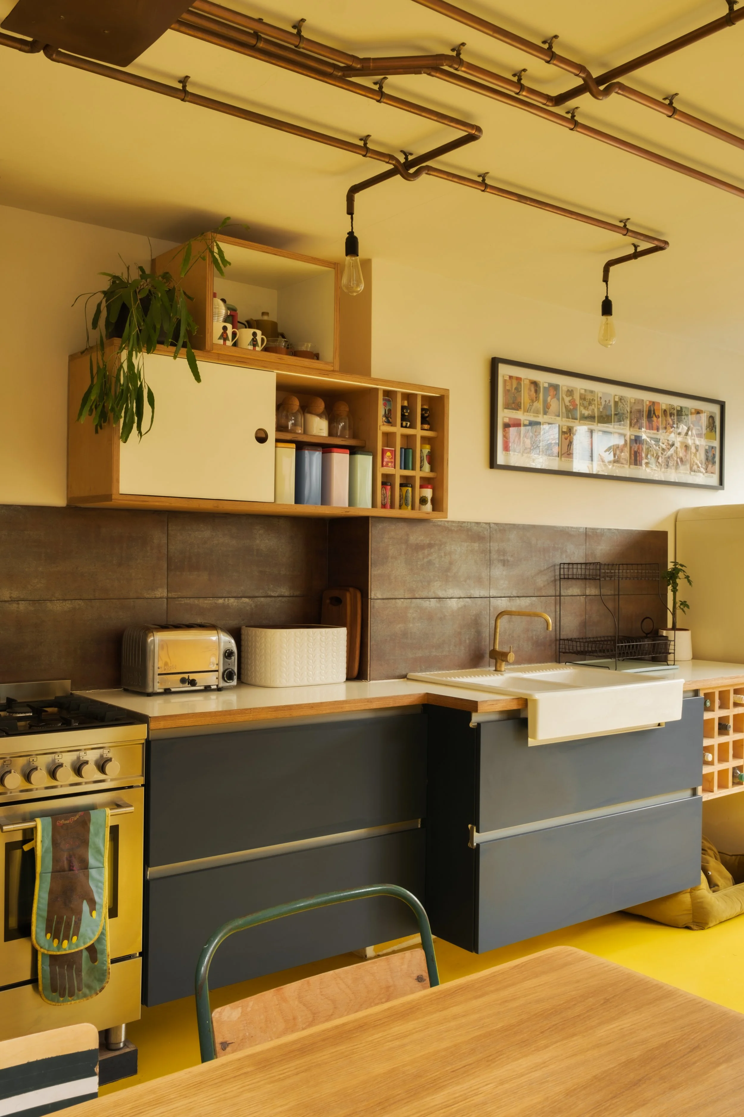 A modern kitchen with dark blue cabinets, a white farmhouse sink, a wooden countertop, and open shelving above. The ceiling features exposed copper piping and light bulbs. A framed collage of comics or art hangs on the wall. Kitchen appliances includ