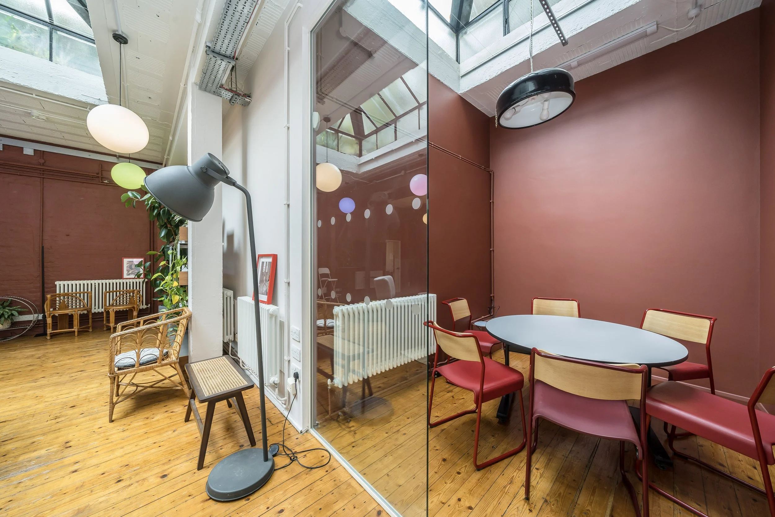 Interior of a modern cafe with wooden floors, pink walls, and colorful hanging light bulbs, featuring a glass partition and a dining table with red and beige chairs.