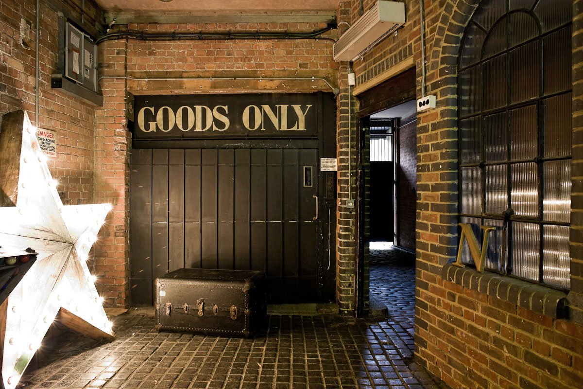 Industrial brick wall hallway with a black sliding door labeled 'GOODS ONLY' and a vintage dark suitcase in front of it. A lit wooden star decoration is on the left, and a window with metal framing and a 'N' letter on the right.