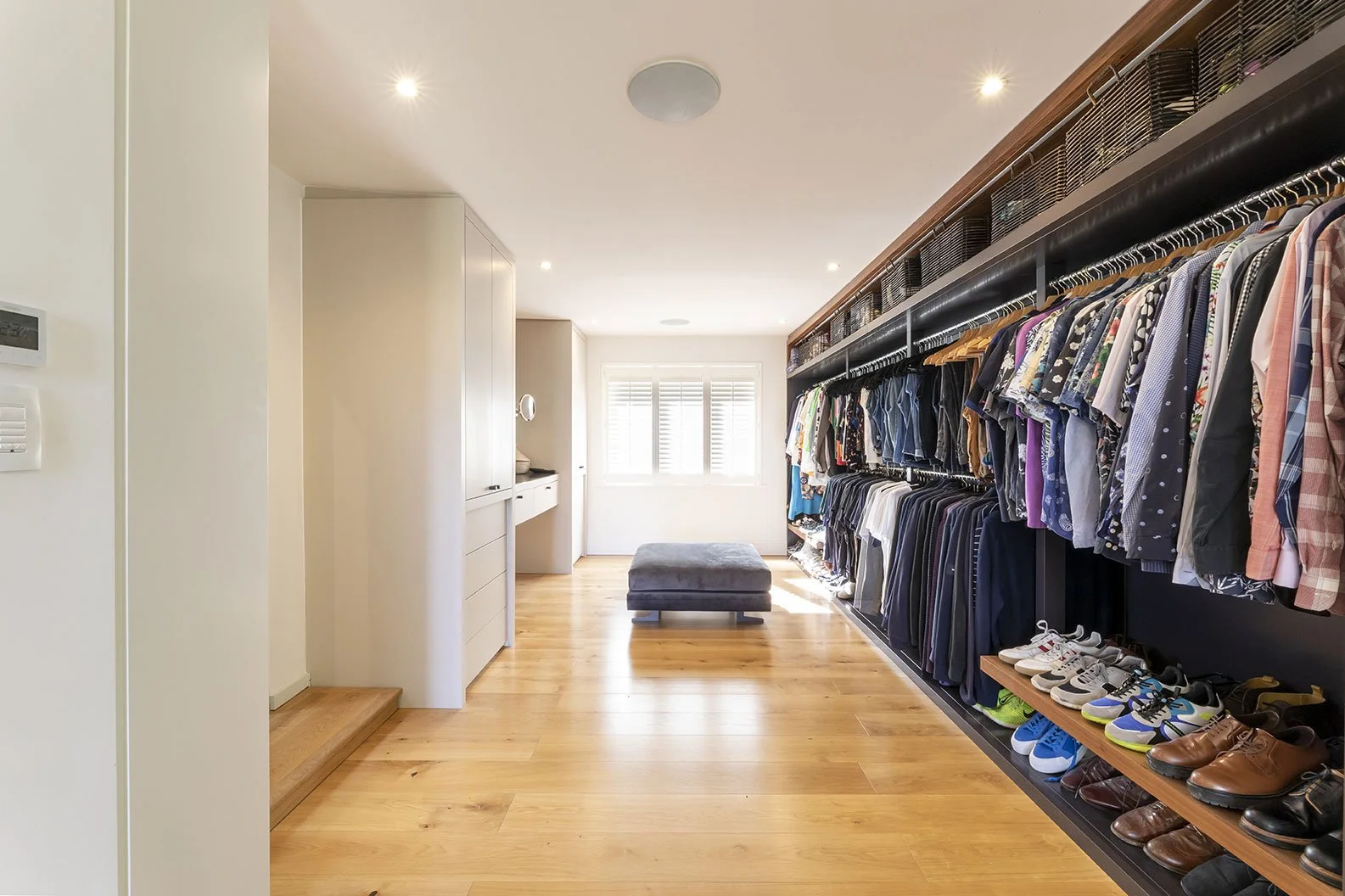 A spacious walk-in closet with wooden flooring, white walls, a large window with blinds, and clothes hanging on a black rack. Shoes are lined up on a wooden shelf below the hanging clothes. There is a grey ottoman in the middle of the room.