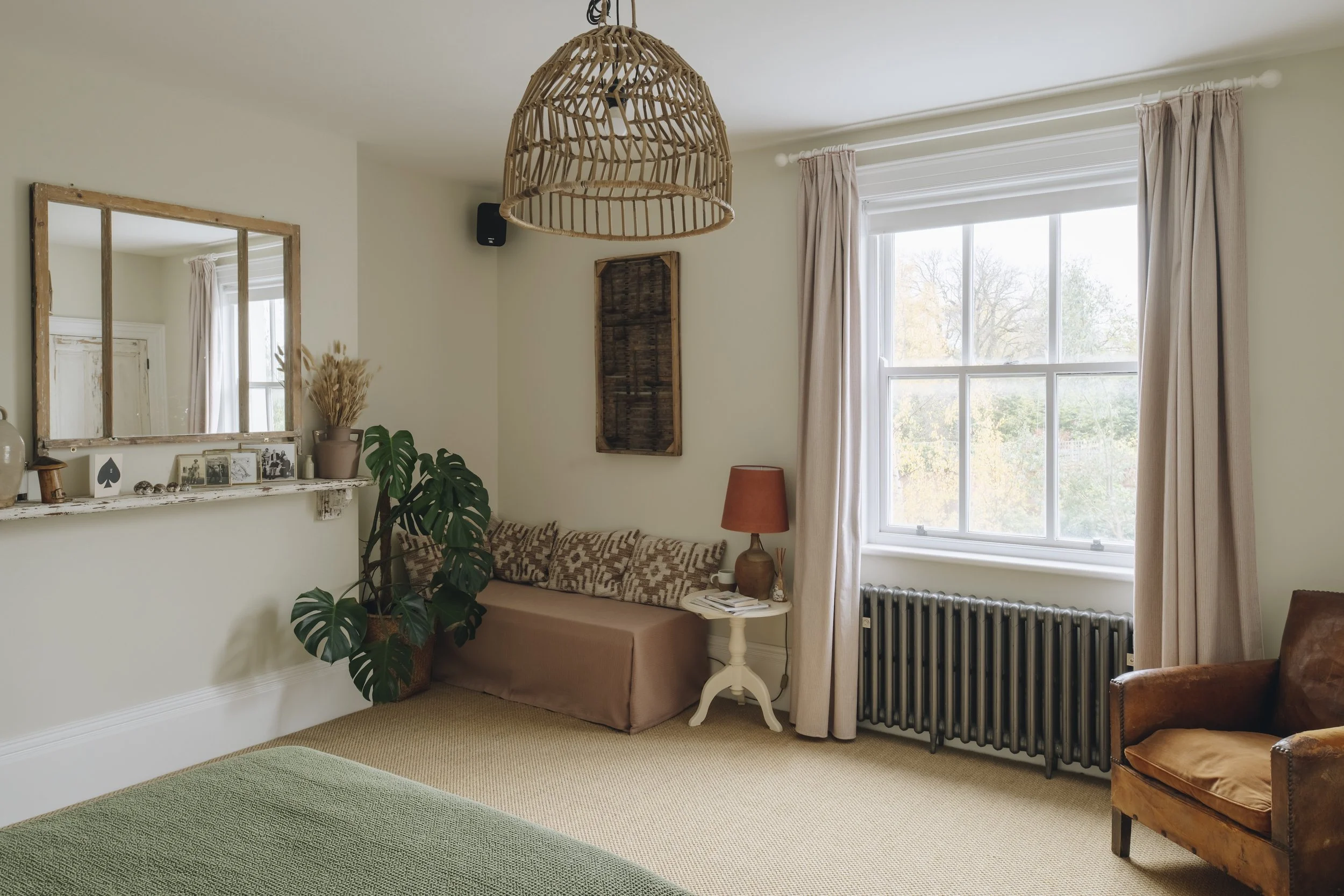 A cozy living room with a large window, light-colored walls, and minimal decor. There is a mirror, plants, a couch with patterned cushions, a side table with a lamp, and an armchair. The room has a wicker light fixture hanging from the ceiling and be