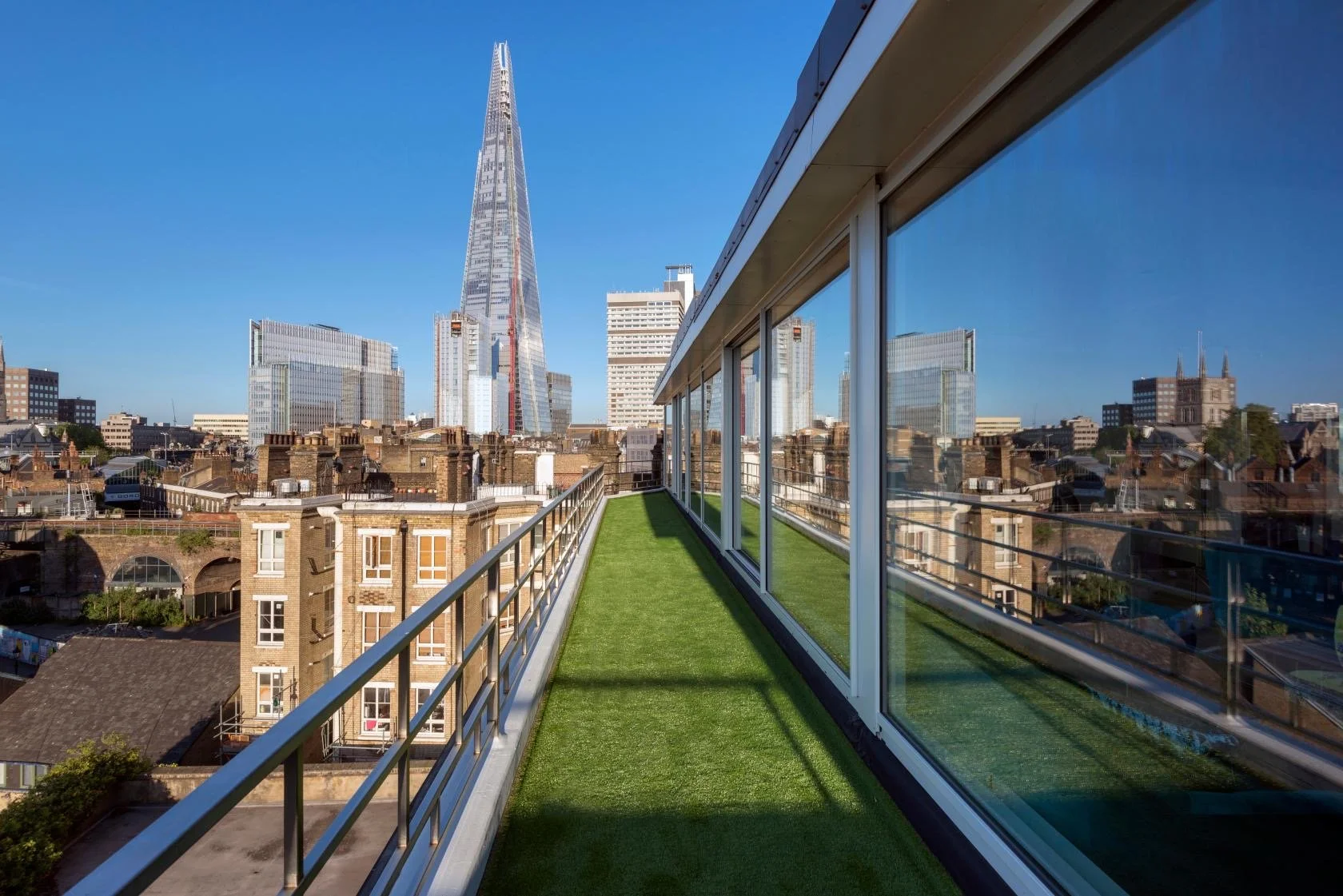 View of London city skyline from a rooftop balcony with artificial grass and glass walls, featuring the Shard skyscraper in the background.