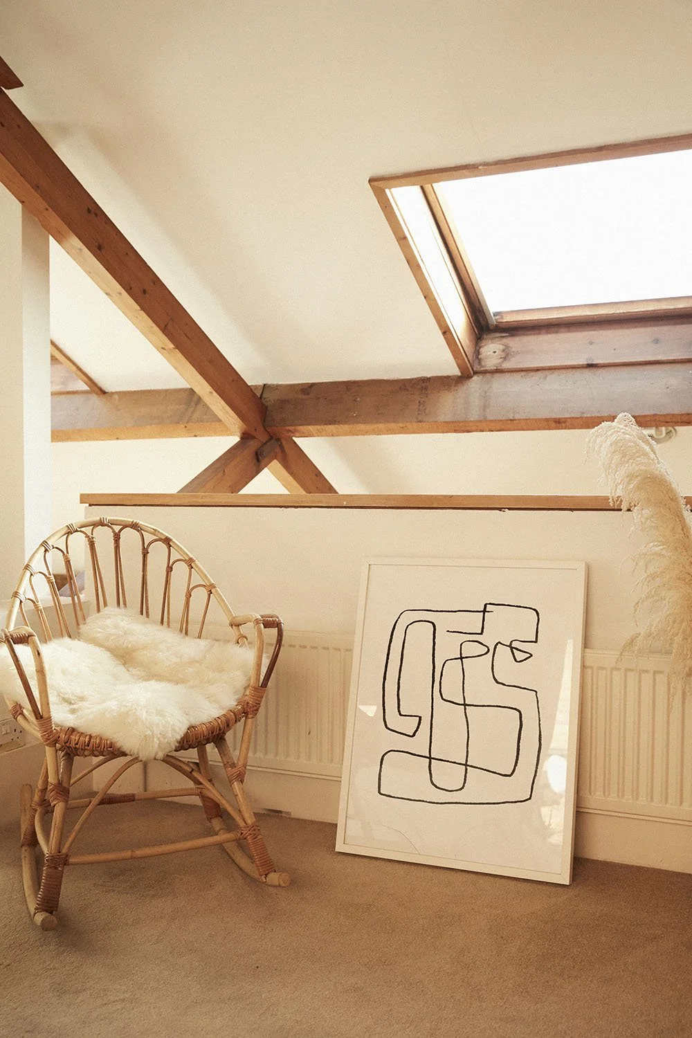 A cozy corner featuring a wooden rocking chair with a white furry cushion, a large abstract line drawing leaning against the wall, and a tall pampas grass plant on the right, all under a skylight in a room with wooden beams.