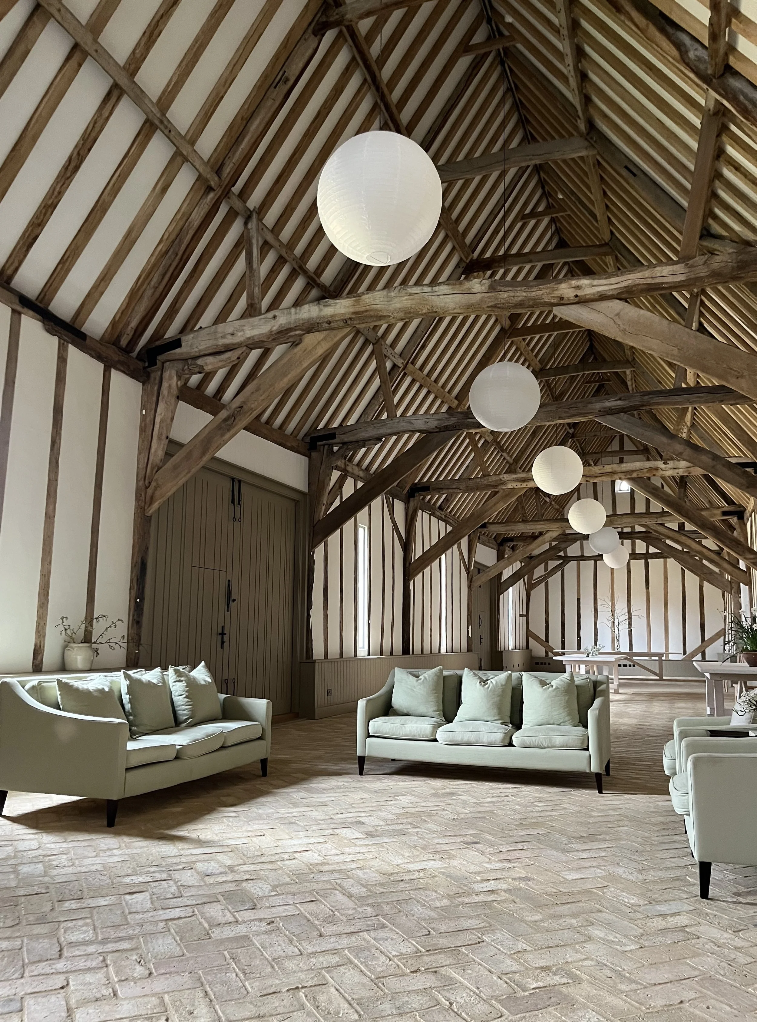 A spacious interior with a high vaulted wooden ceiling supported by rustic beams, white paper lantern pendant lights hanging from the ceiling, and a sitting area with light green sofas and pillows on a brick floor.