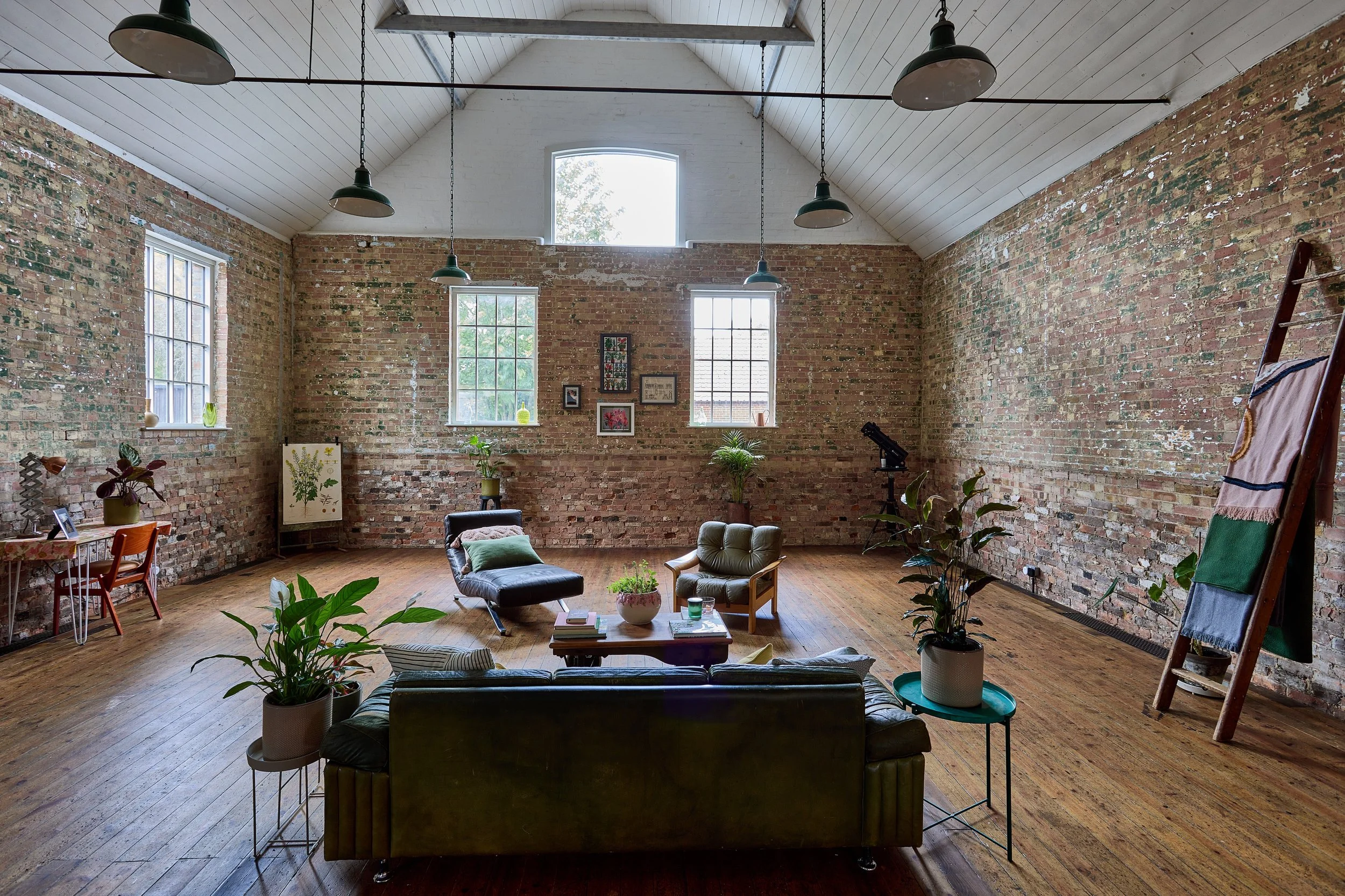 Spacious loft with brick walls, wooden floors, large windows, various plants, a sitting area with a leather sofa, armchair, and coffee table, art on walls, a telescope, and a ladder with hanging fabric.