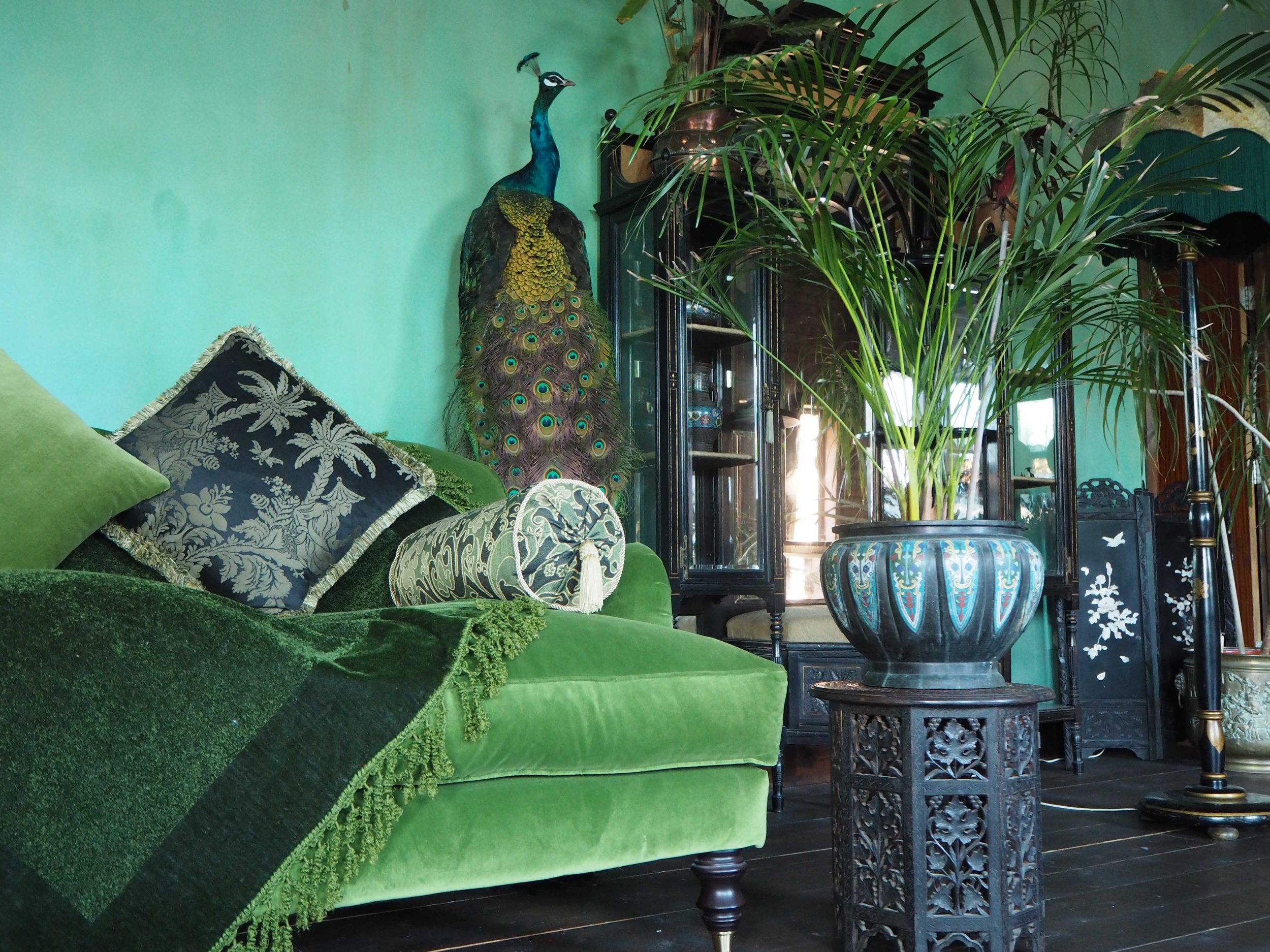 A living room corner with a green velvet sofa adorned with patterned pillows and a tasseled bolster. Behind the sofa, a large peacock sculpture stands outdoors near the window, surrounded by green plants in decorative pots. Sunlight filters through t