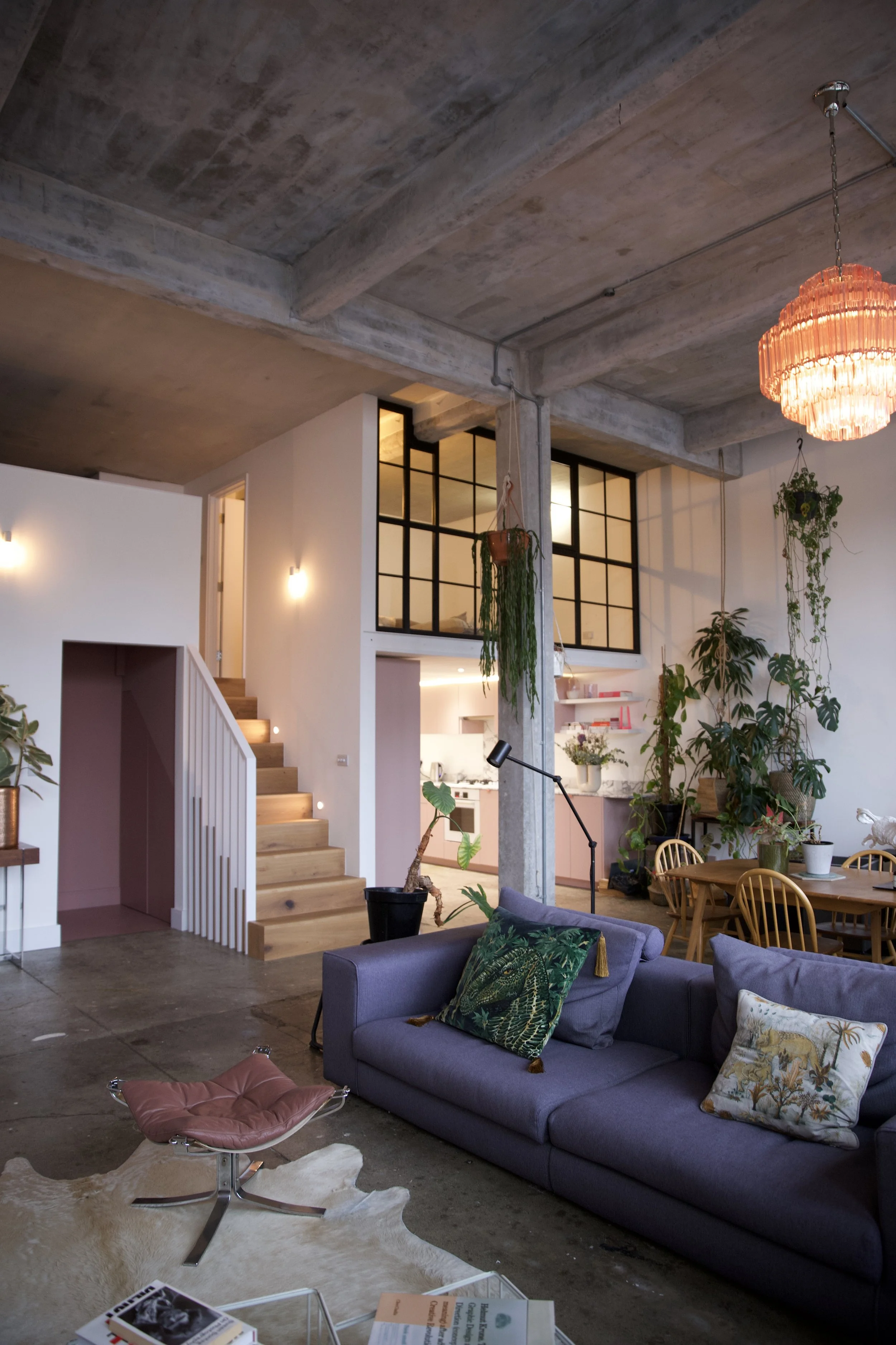 Interior view of a modern, industrial-style living room with a concrete ceiling, wooden staircase, purple sofa with decorative pillows, hanging plants, and a dining area with wooden chairs. Kitchen is visible in the background.
