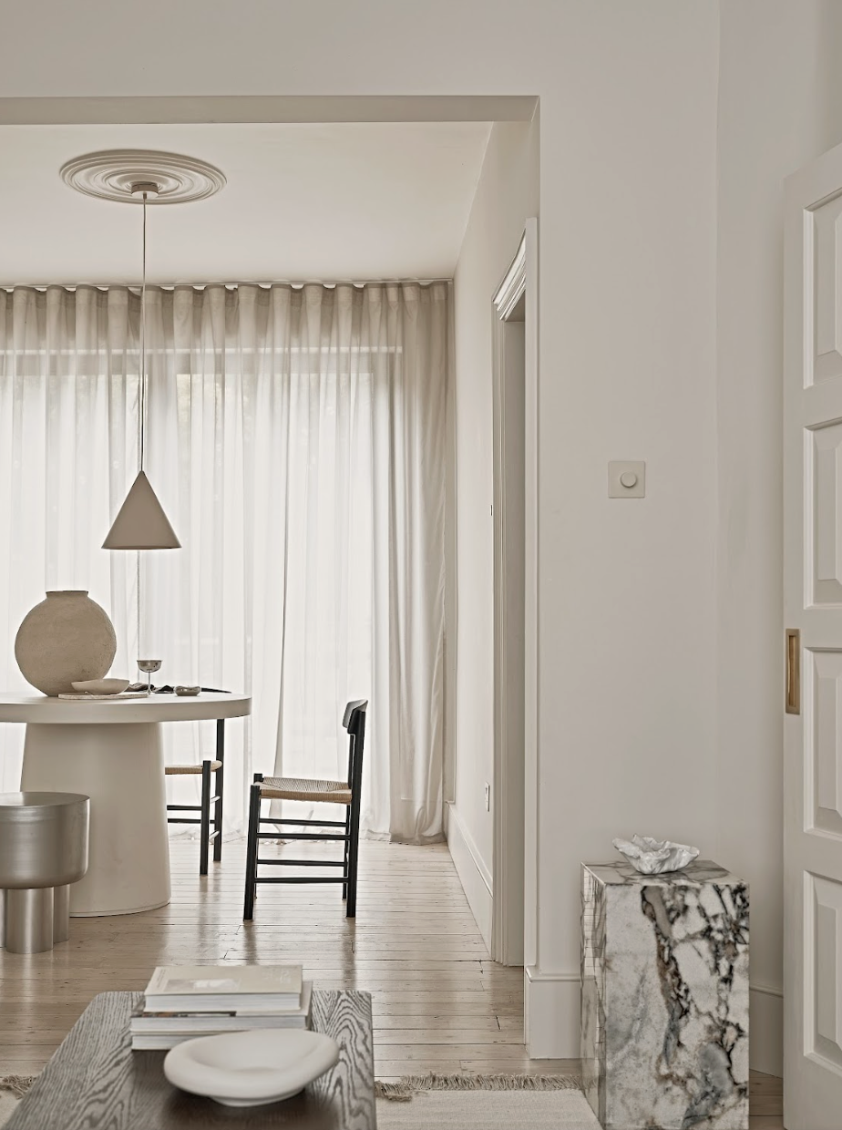 A minimalist, light-colored dining area with a round white table, black chairs, and a curtain-covered window in the background. Decor includes a large rounded vase, a small bowl, and a wine glass on the table. A pendant lamp hangs above the table, an