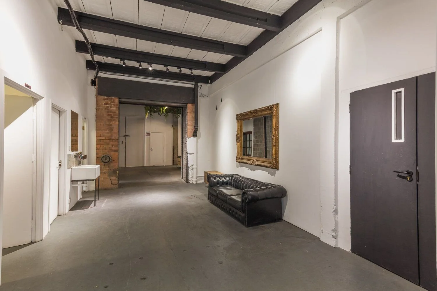 Interior of an industrial-style building with exposed brick and white walls, black ceiling beams, a black leather couch, a large ornate gold mirror, and a gray door.