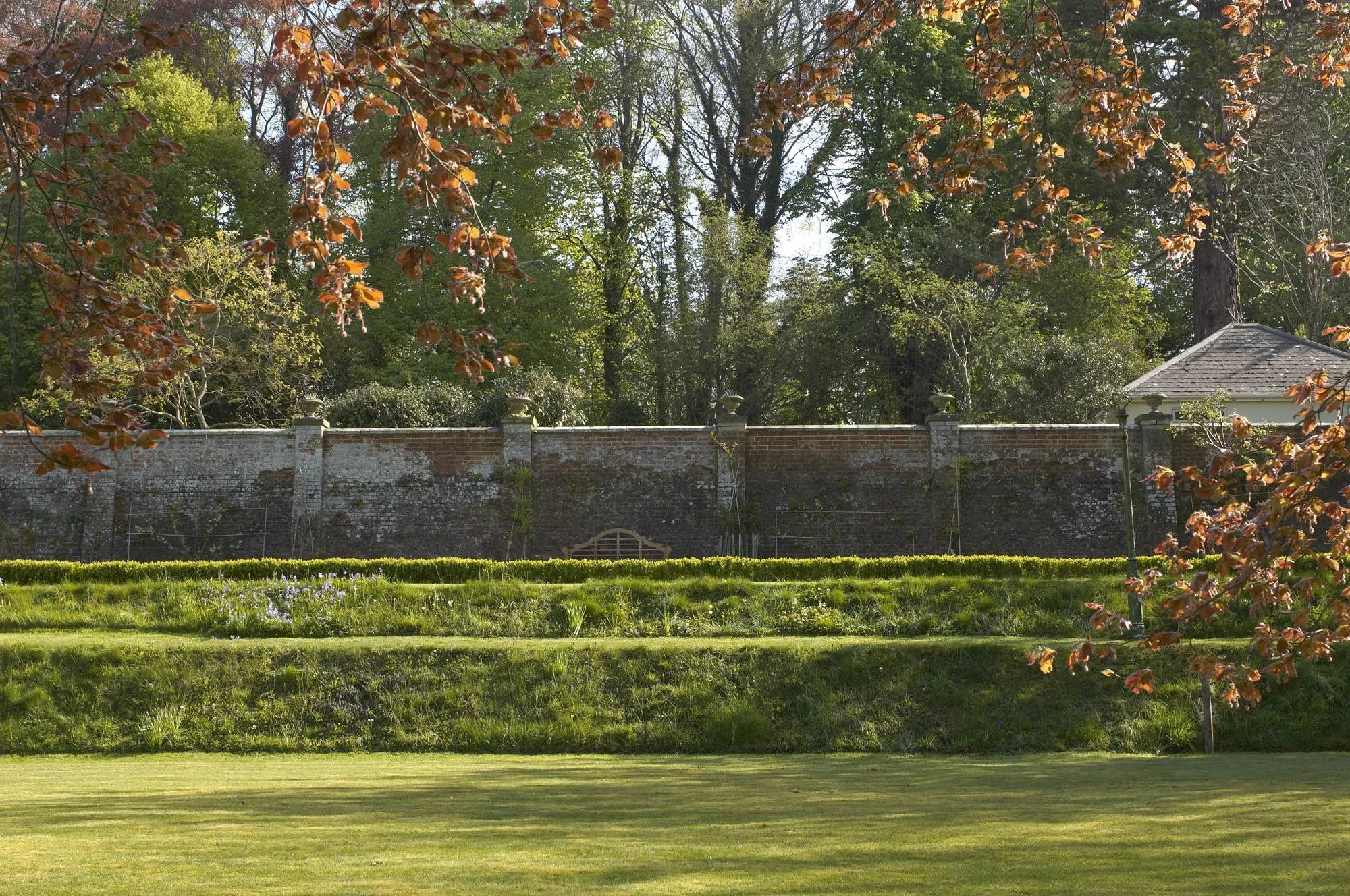 A well-maintained green lawn with tiered grass sections, a brick wall with stone columns in the background, and a gazebo on the right side surrounded by trees with green and orange leaves.