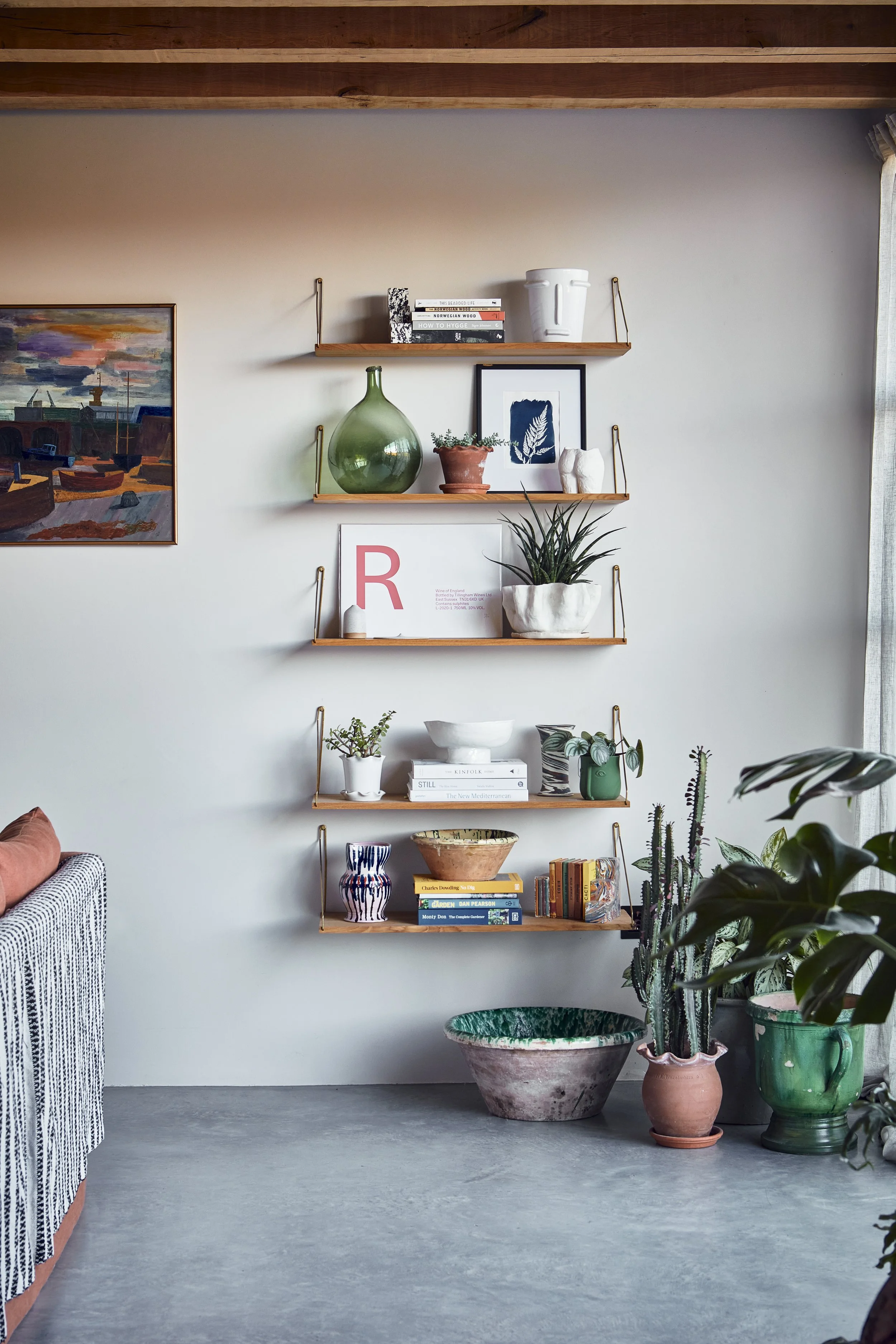 Decorative living room wall with three wooden shelves holding plants, books, framed artwork, and ceramics, with potted plants on the floor.
