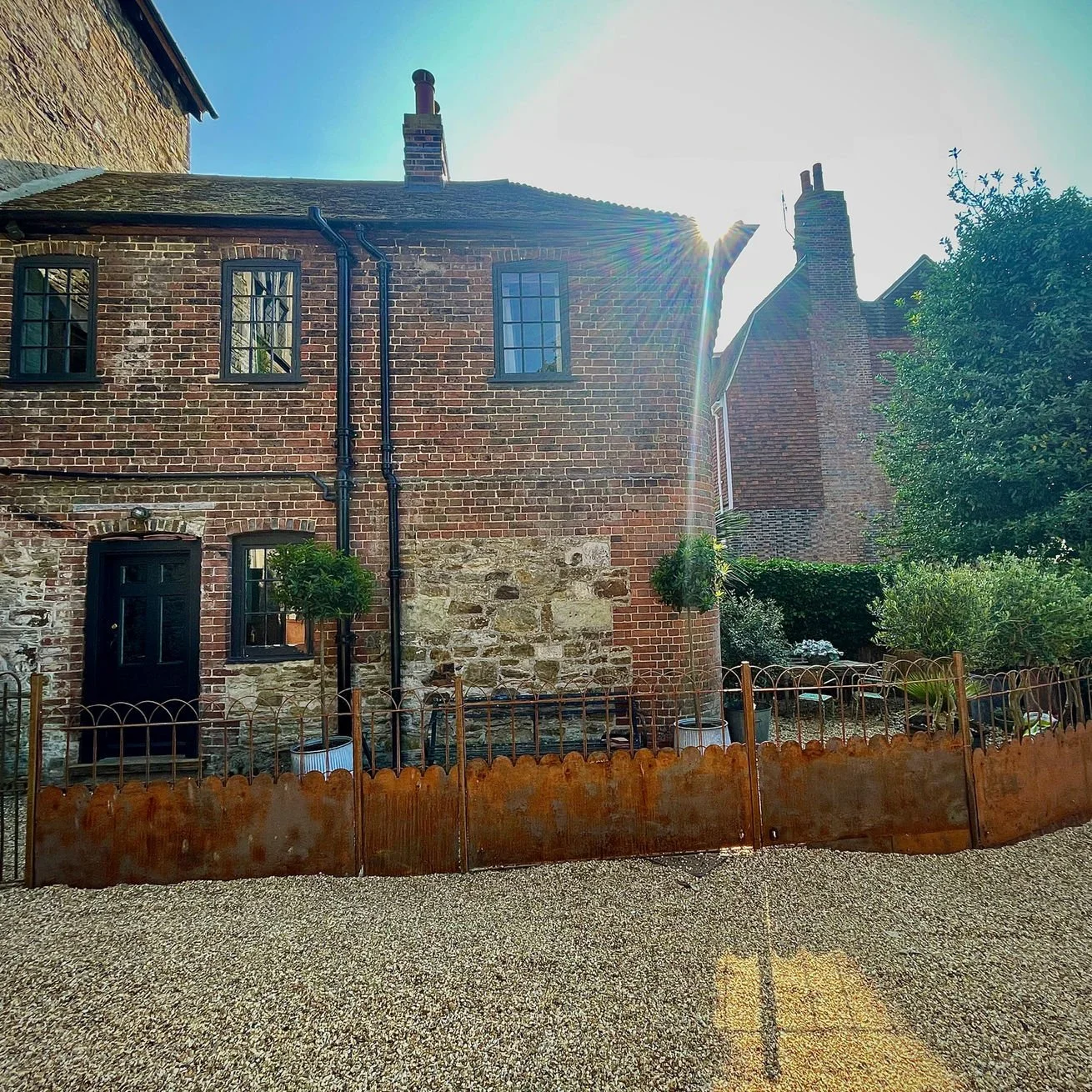 A brick house with black-framed windows, a black door, and a small garden with trees, shrubs, and a decorative fence, under a bright sun in a clear sky.