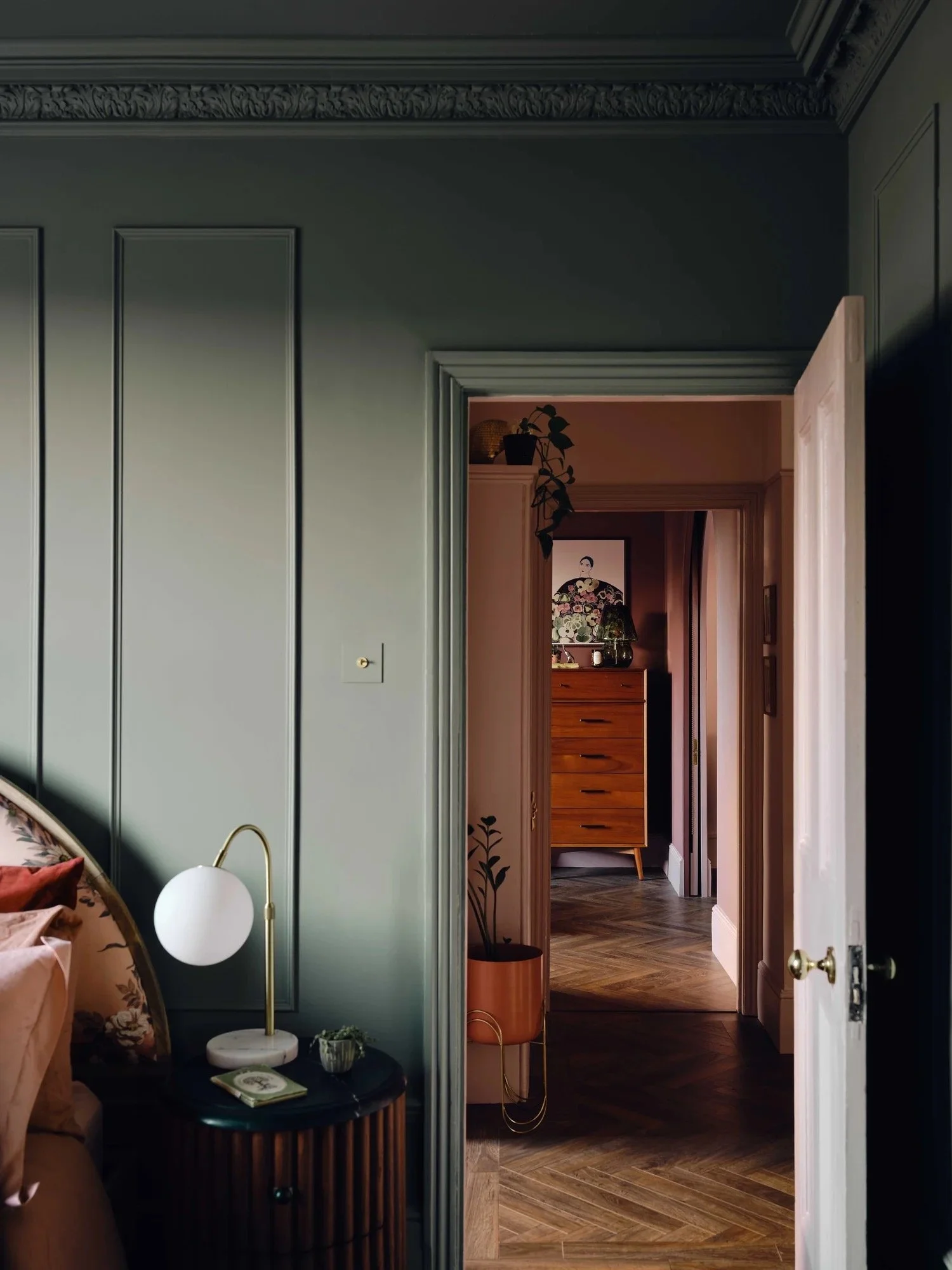 Interior view of a bedroom with dark green wall, side table with a white lamp and books, draped fabric on the bed, and a doorway leading to a hallway with wooden furniture and potted plants.