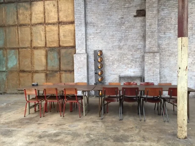Industrial-style room with wooden tables and red chairs, exposed brick wall, large rusty metal door, a set of stage lights against the wall, and a concrete floor.