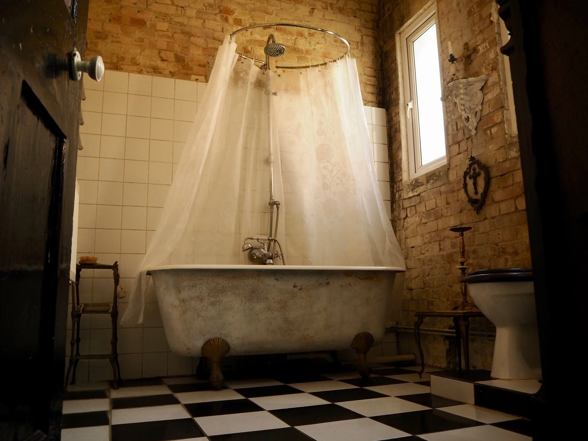 A rustic bathroom with exposed brick walls, a vintage clawfoot bathtub partially covered by a sheer shower curtain, and a checkered black and white tile floor. There is a small window, decorative wall ornaments, and an old-fashioned pedestal sink.