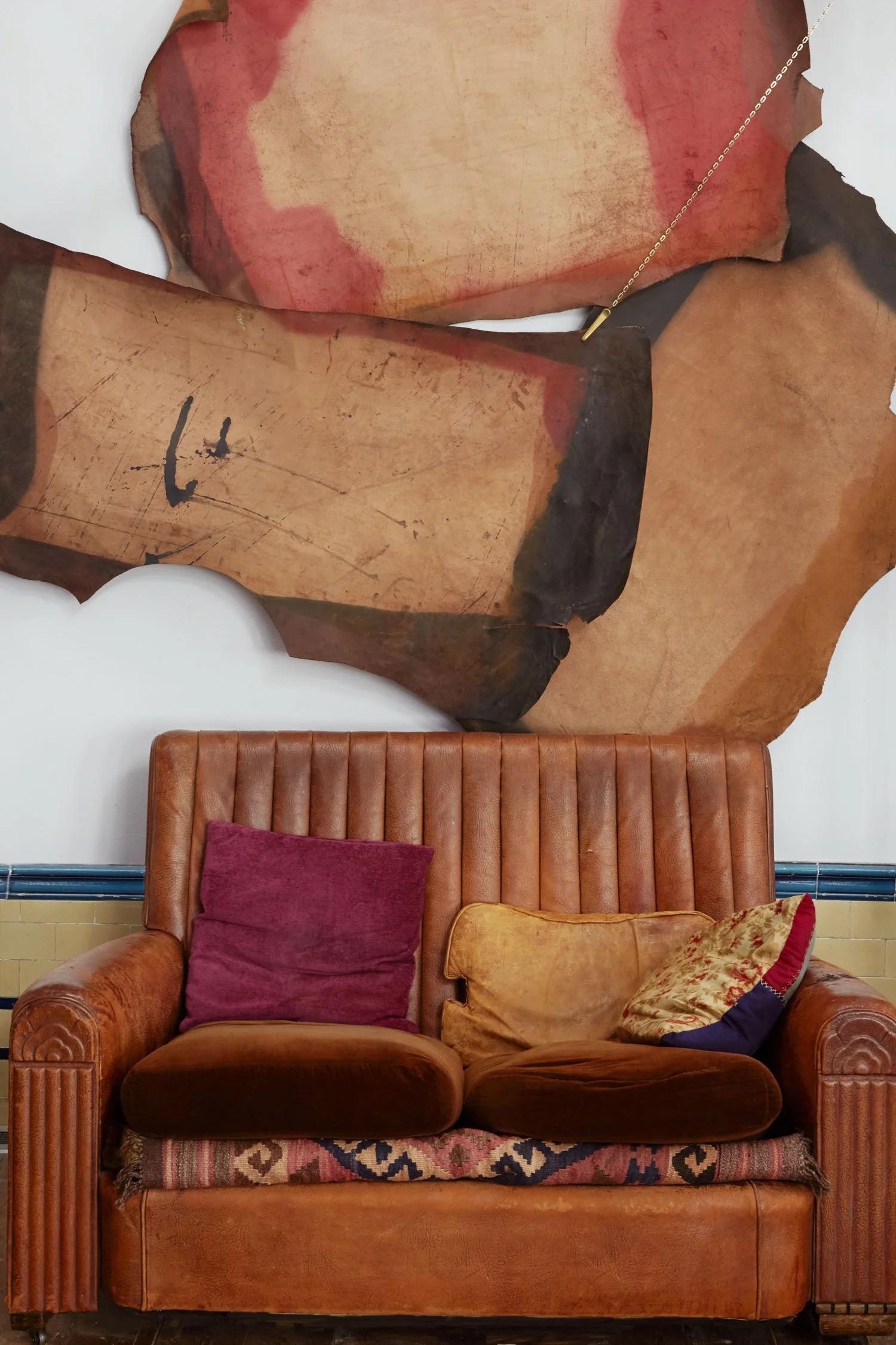 A vintage orange leather sofa with three assorted cushions in front of a white wall. Above the sofa, there is an abstract artwork with torn paper textures in shades of red, orange, black, and beige.