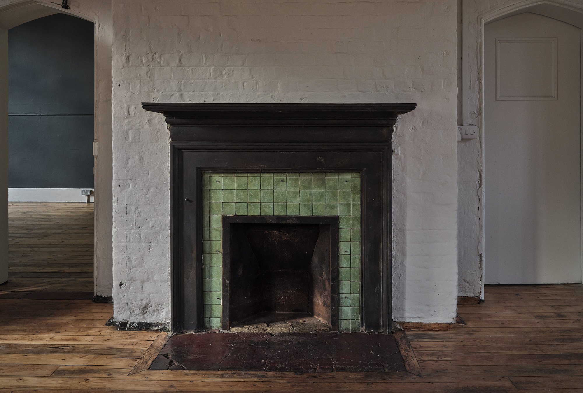 Old fireplace with black mantel and green tiled surround in a white brick wall, on a wooden floor.