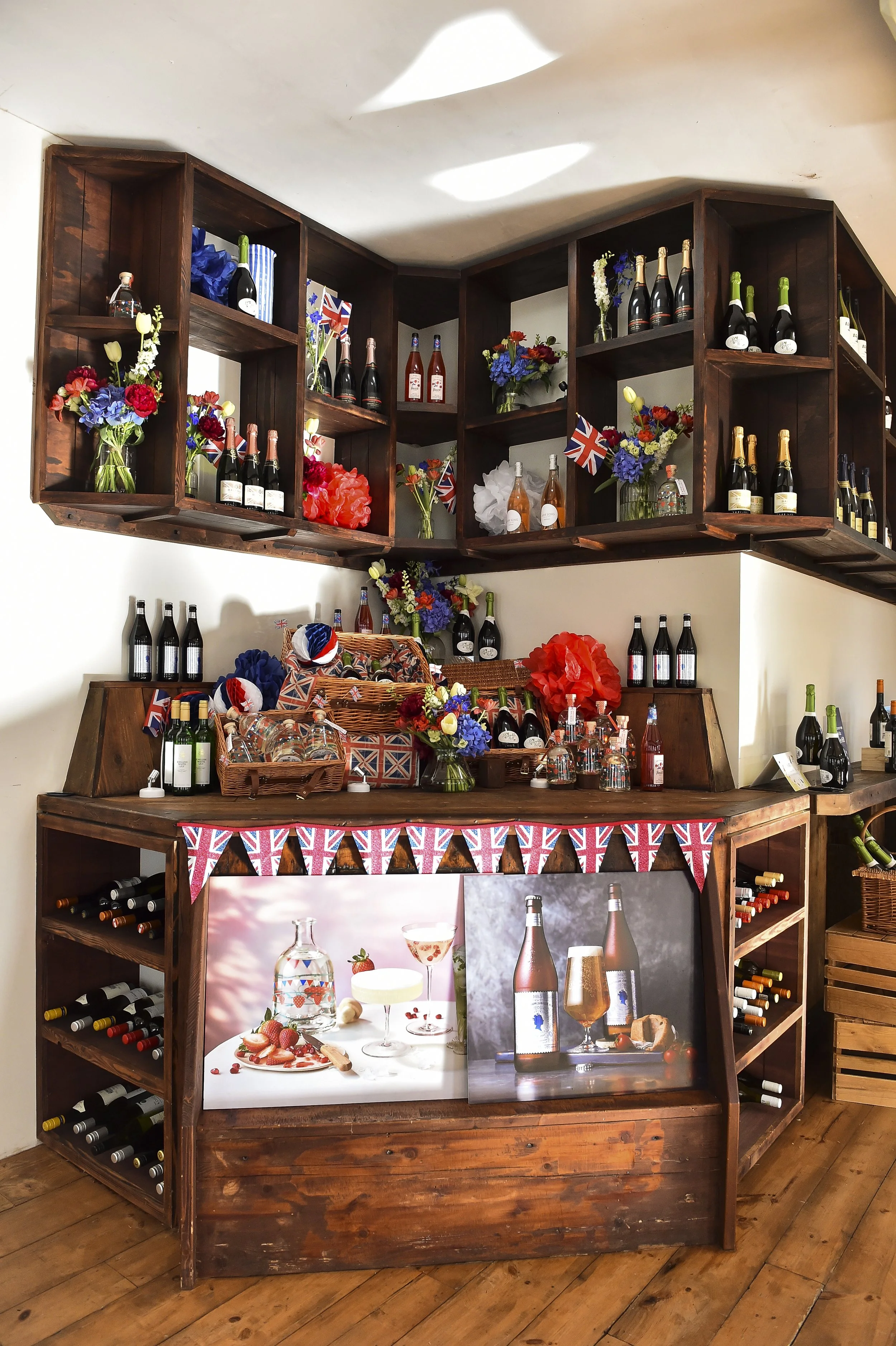 Display of various bottles of wine and champagne, floral arrangements, Union Jack decorations, and images of cocktails and beverages on a wooden bar counter with wine racks on sides.
