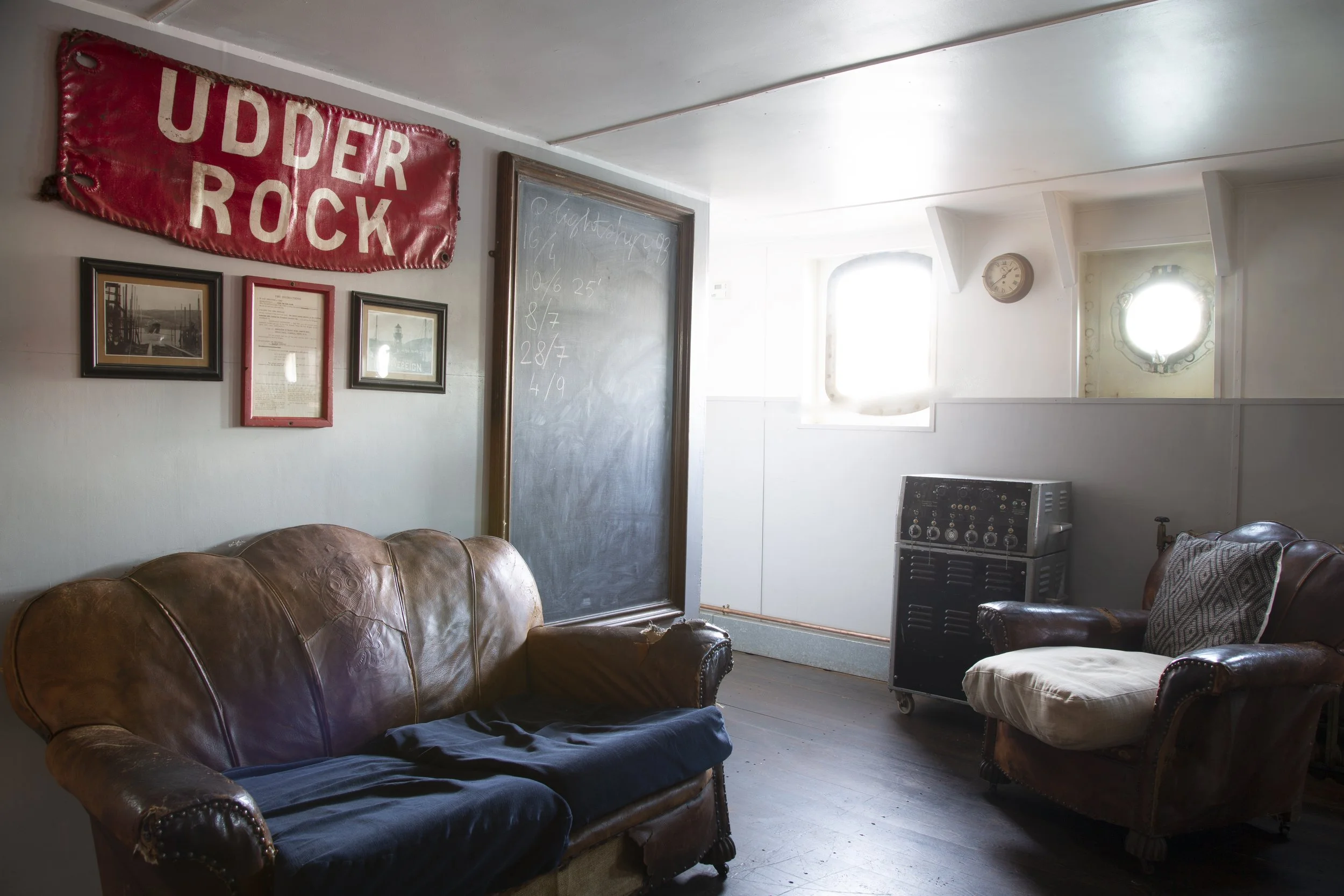 A cozy lounge area featuring a brown leather sofa covered with a dark cloth, an armchair with a patterned pillow, a vintage black refrigerator, and a double window with sunlight. The wall is decorated with framed pictures and a red sign that reads 'U