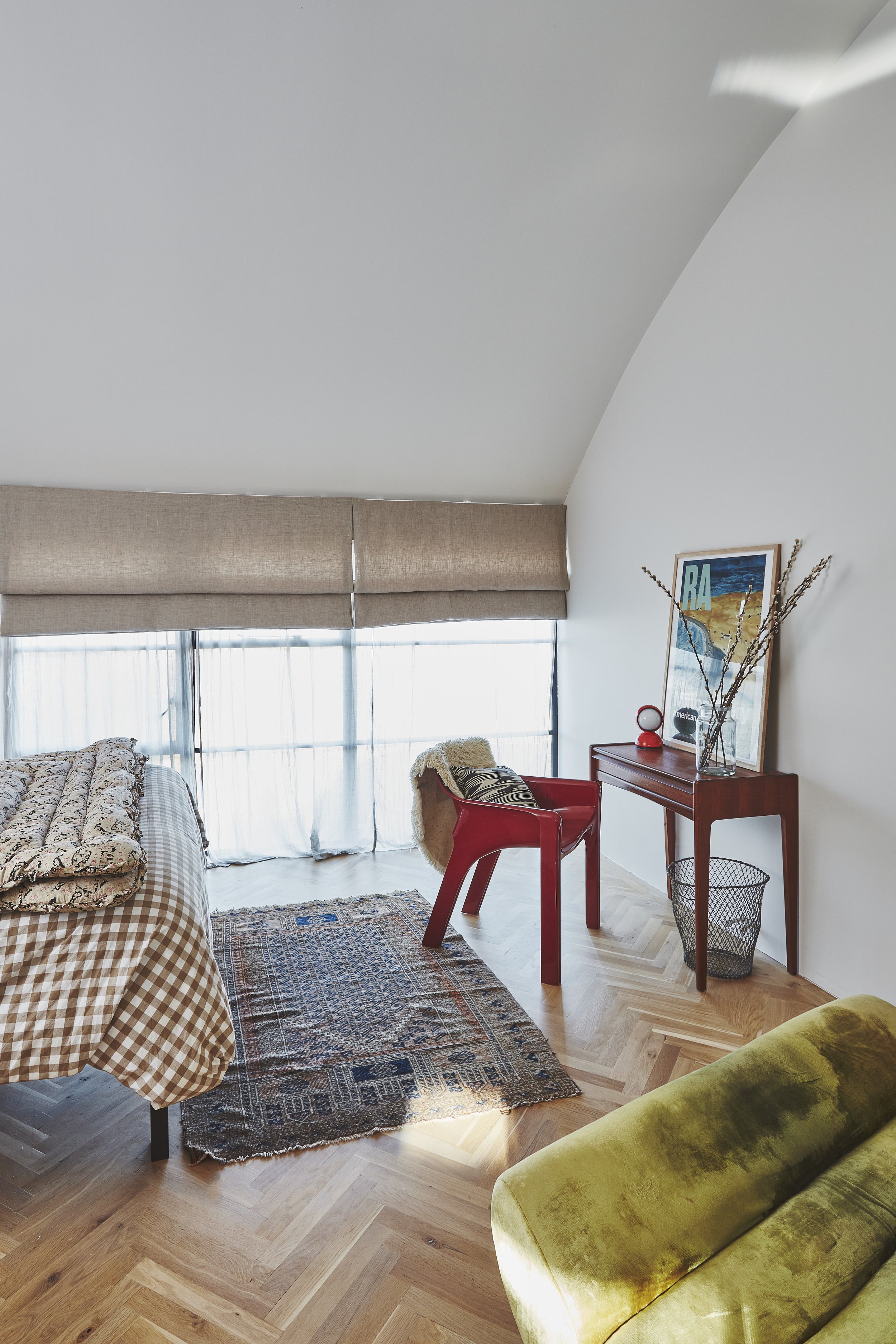 A cozy living room with a large window covered by beige blinds, a patterned sofa with pillows, a red chair with a sheepskin throw, a wooden side table with a framed poster, a vase with dried branches, and a green velvet sofa at the bottom of the imag