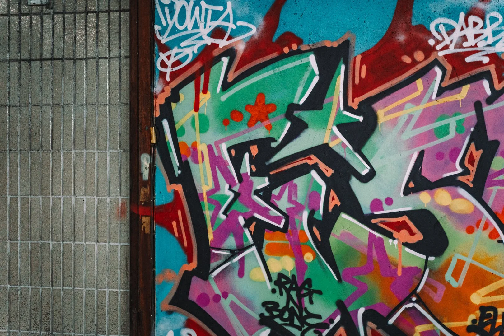 Colorful graffiti art on a wall featuring abstract shapes, bright colors, and spray paint tags. The left side of the image shows a gray brick wall, and the right side showcases multicolored spray-painted artwork.