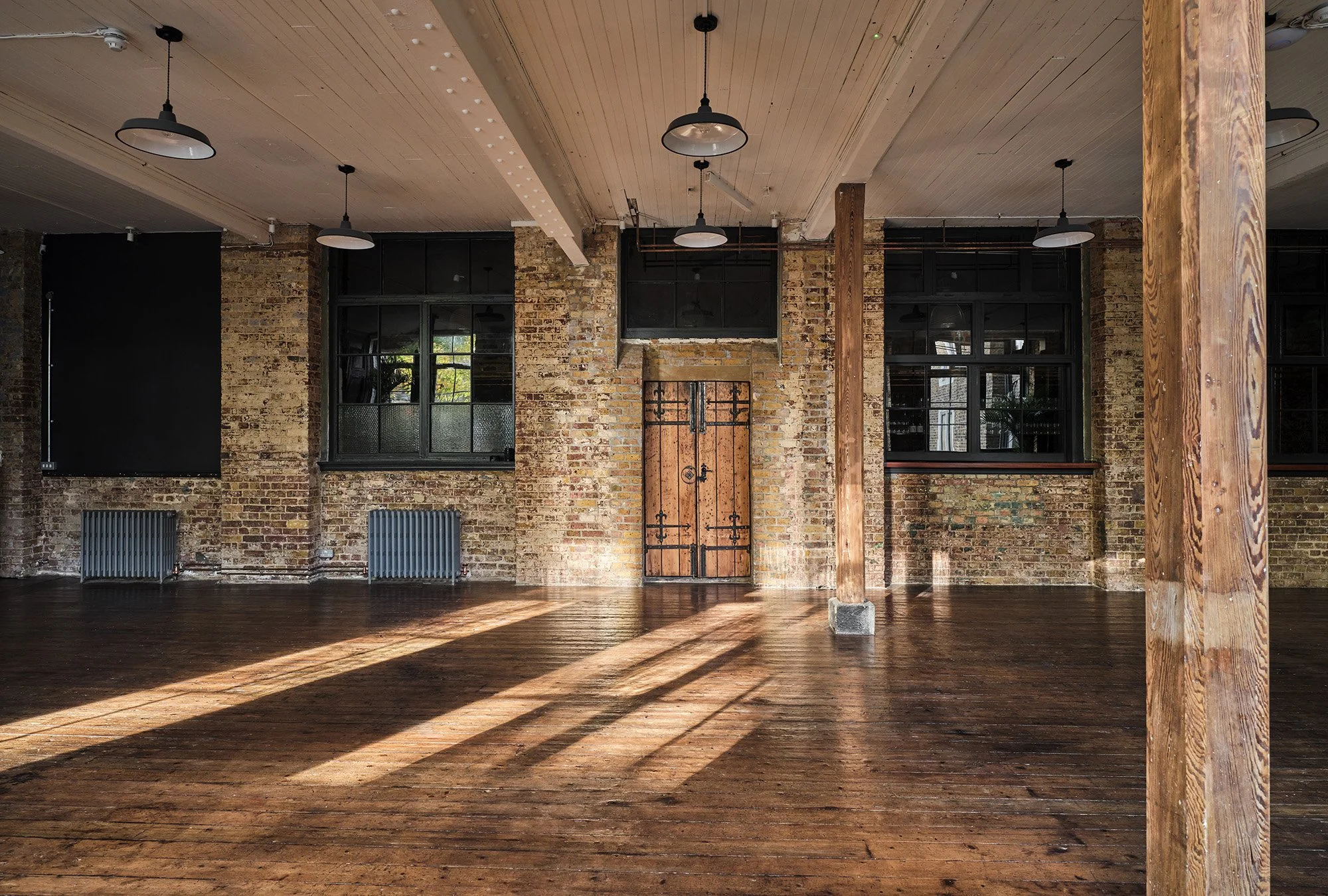 Empty, spacious room with exposed brick walls, wooden floor, large windows, wooden columns, and hanging pendant lights.