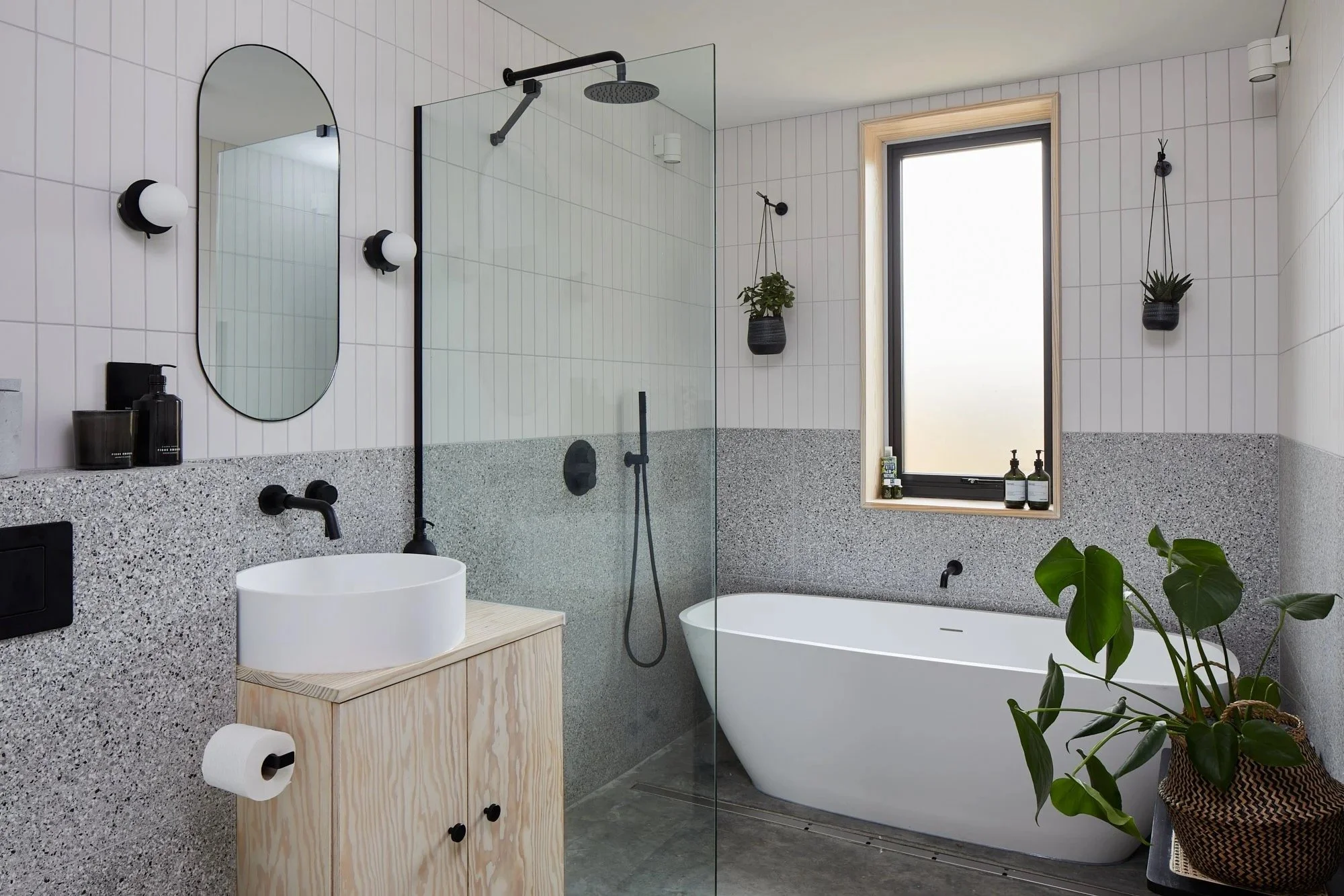 Modern bathroom with white and gray decor, a freestanding bathtub near a window, a small wooden vanity with a round vessel sink, a walk-in shower with a glass partition, black fixtures, and potted plants hanging and on the floor.
