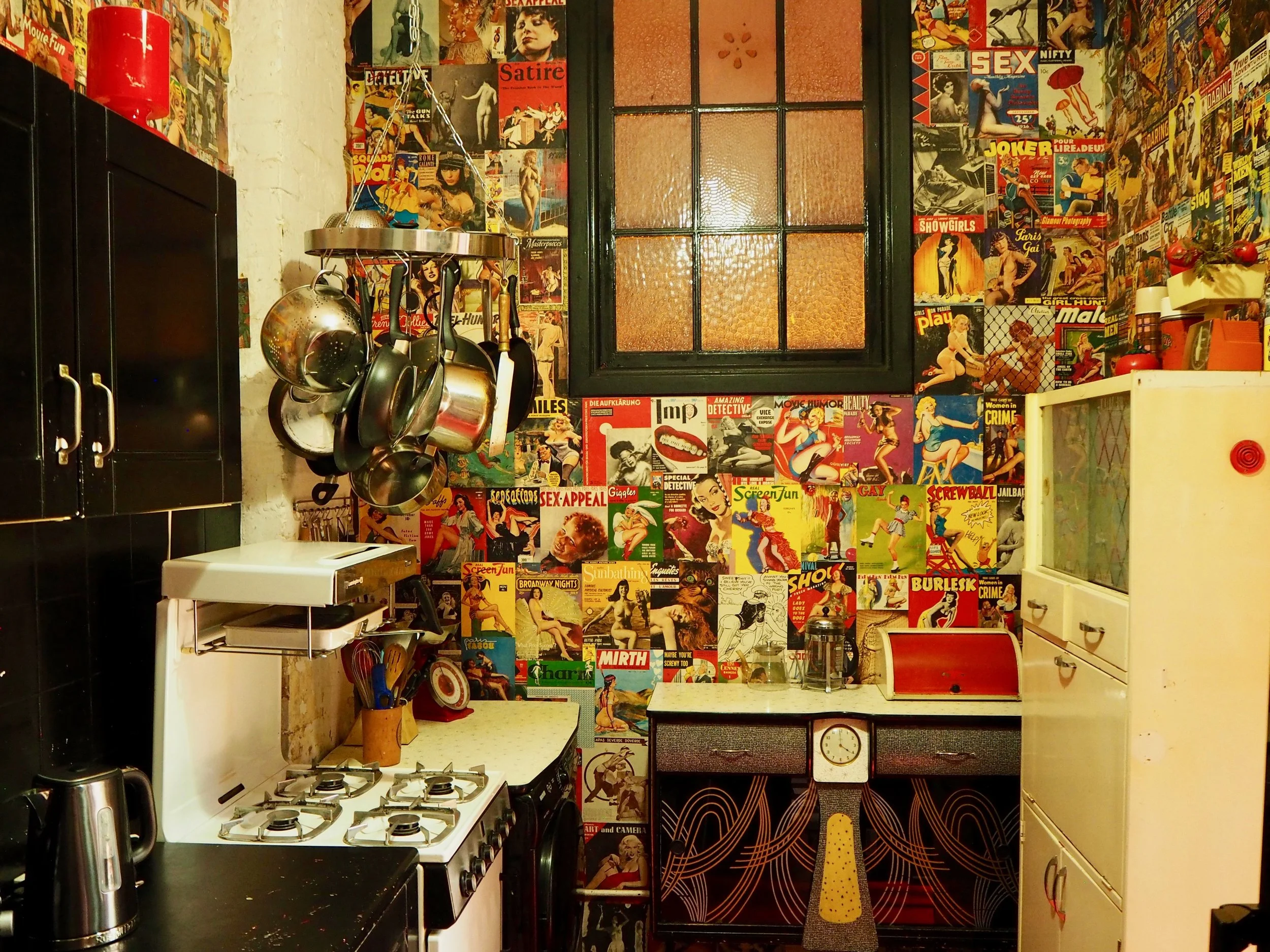 A vintage-style kitchen with a wall covered in colorful comic book covers. The kitchen includes a black cabinet, a white stove, hanging pots and pans, a small white countertop, a white fridge with a red smiley face, and a window with textured glass. 