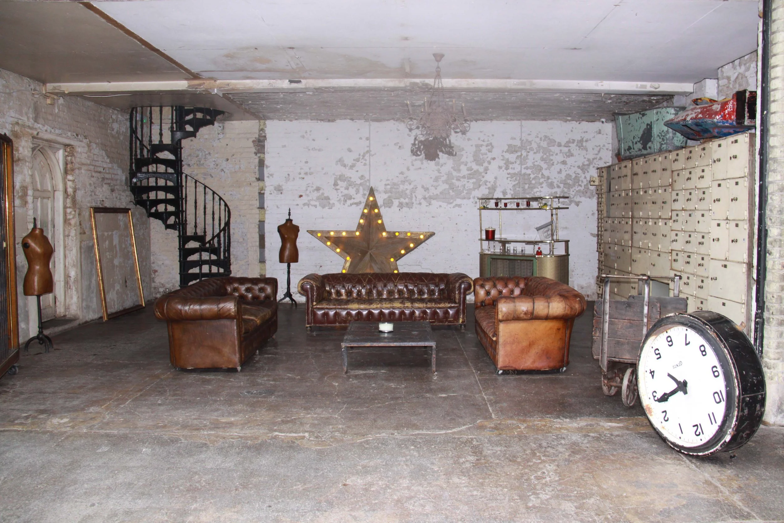 Industrial-style loft interior with three vintage leather sofas, a large illuminated star on the wall, a small coffee table, dress forms, lockers, and a giant clock leaning against the wall.