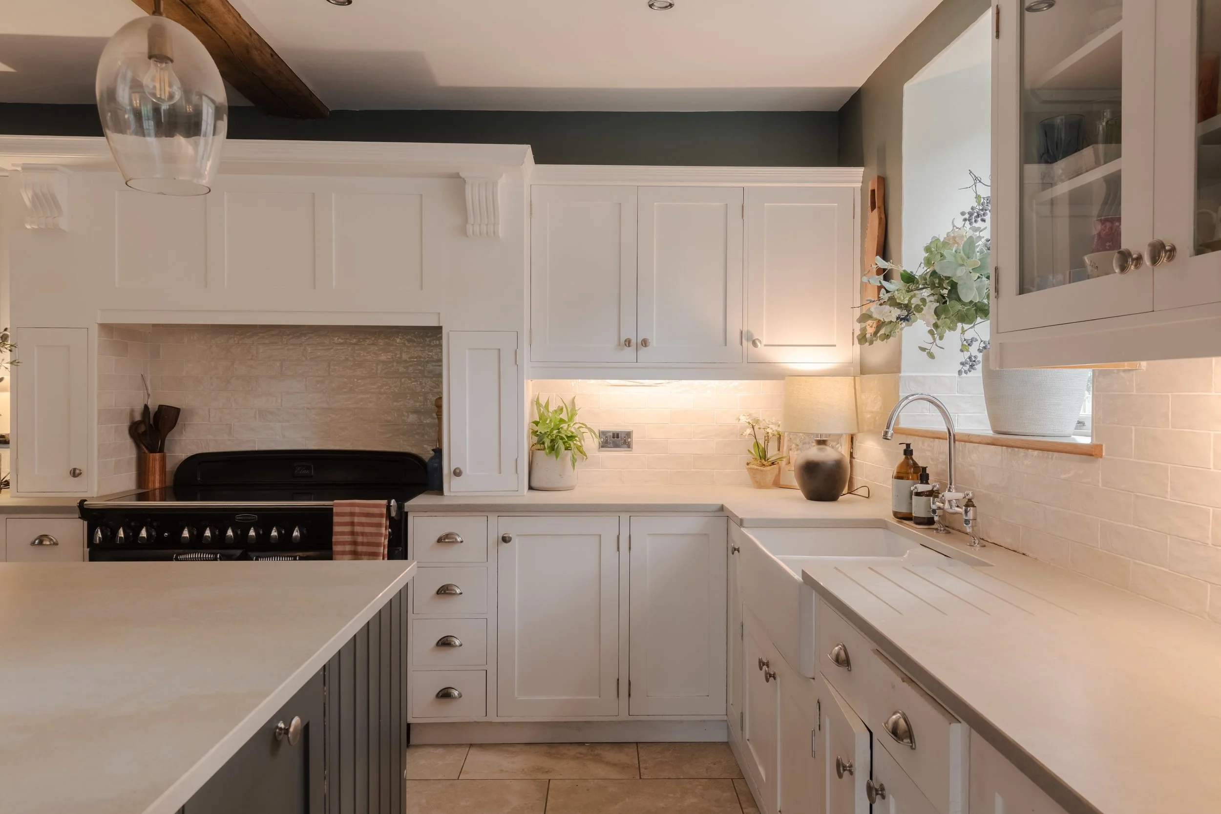 A kitchen with white cabinets, a white brick backsplash, a black stove, and a window with a large potted plant. Items on the counter include a lamp, a plant, soap dispenser, and kitchen utensils.