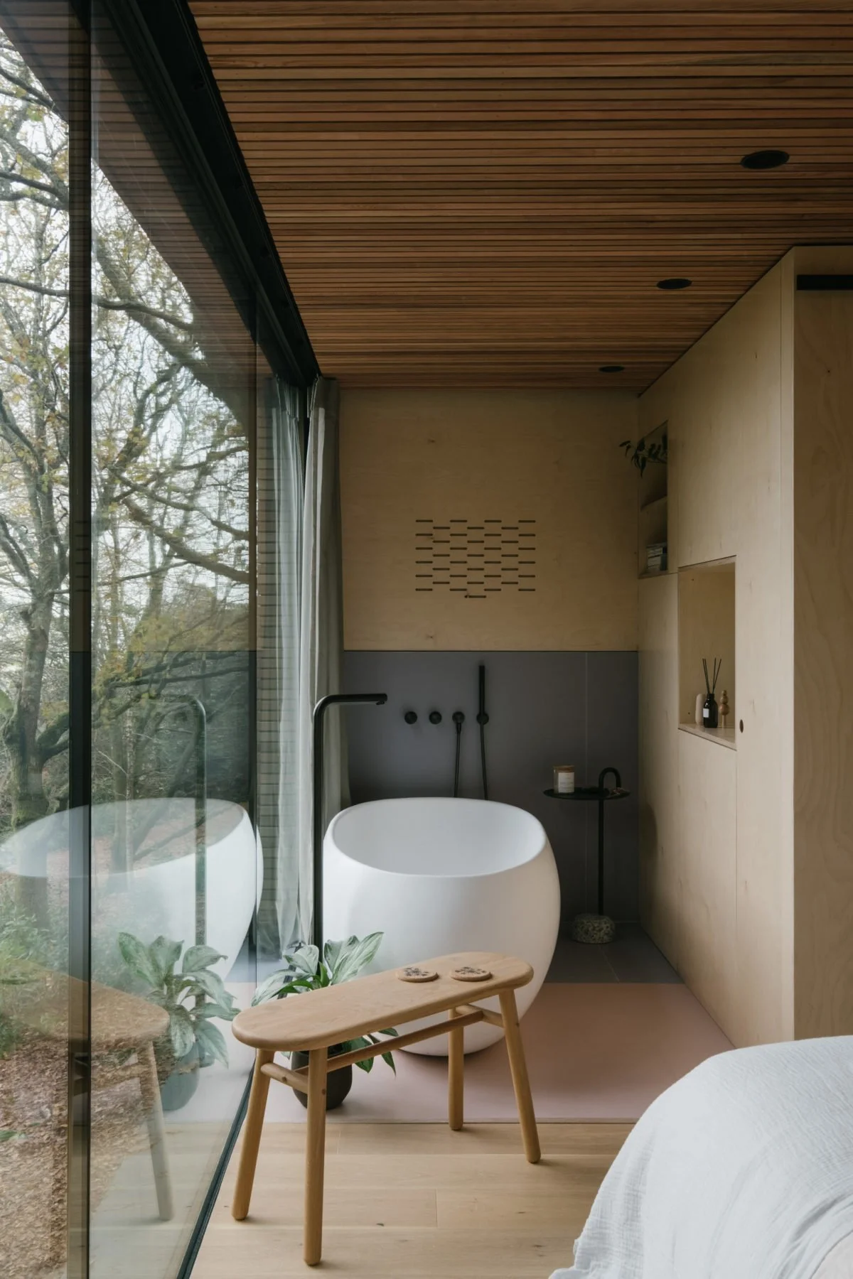 A modern bathroom featuring a freestanding white bathtub next to a large floor-to-ceiling window showing outdoor trees, with a wooden side table, potted plant, and minimalist decor.