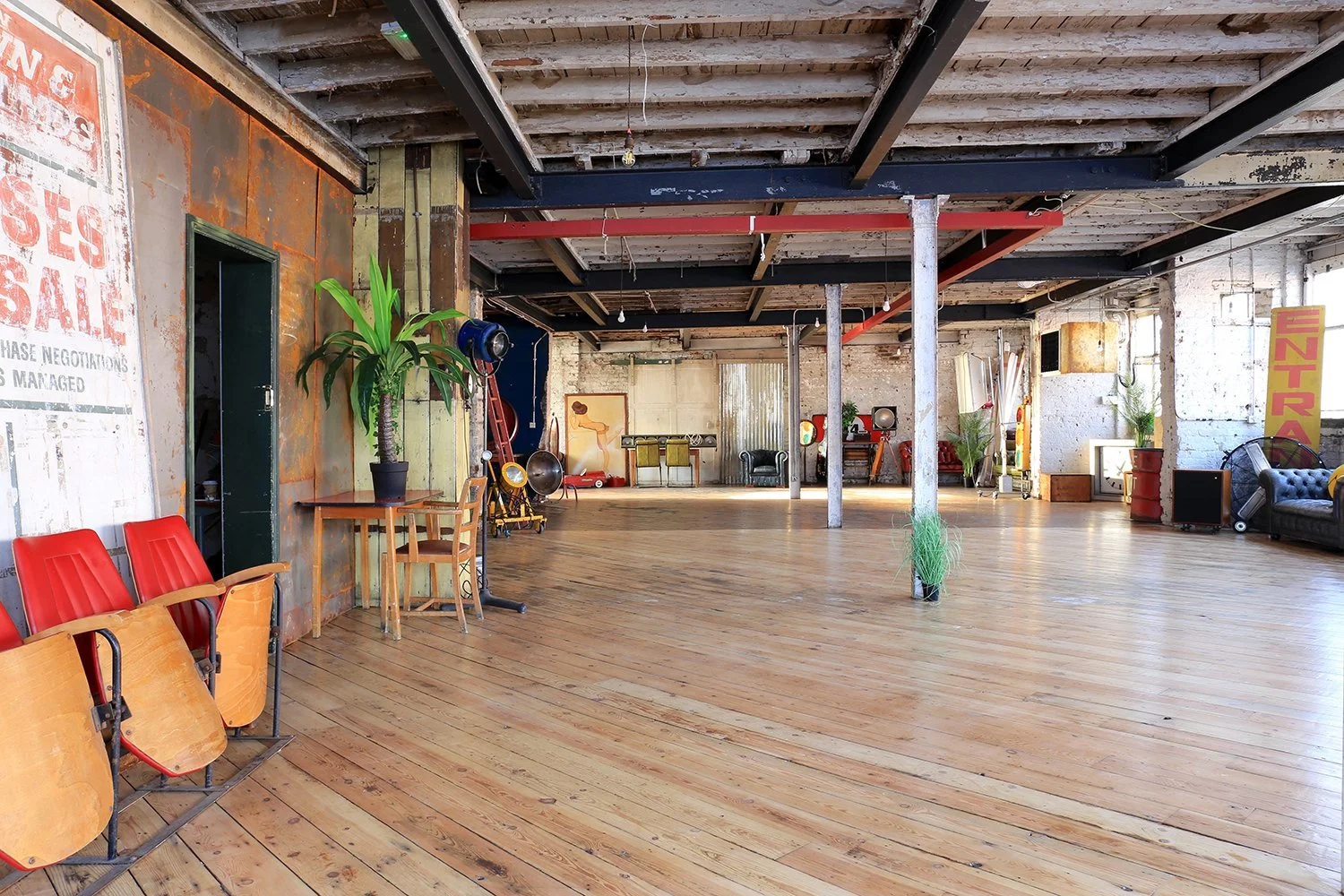 Spacious, industrial-style room with wooden floors, exposed ceiling beams, and with furniture including chairs, sofas, plants, and vintage decor.