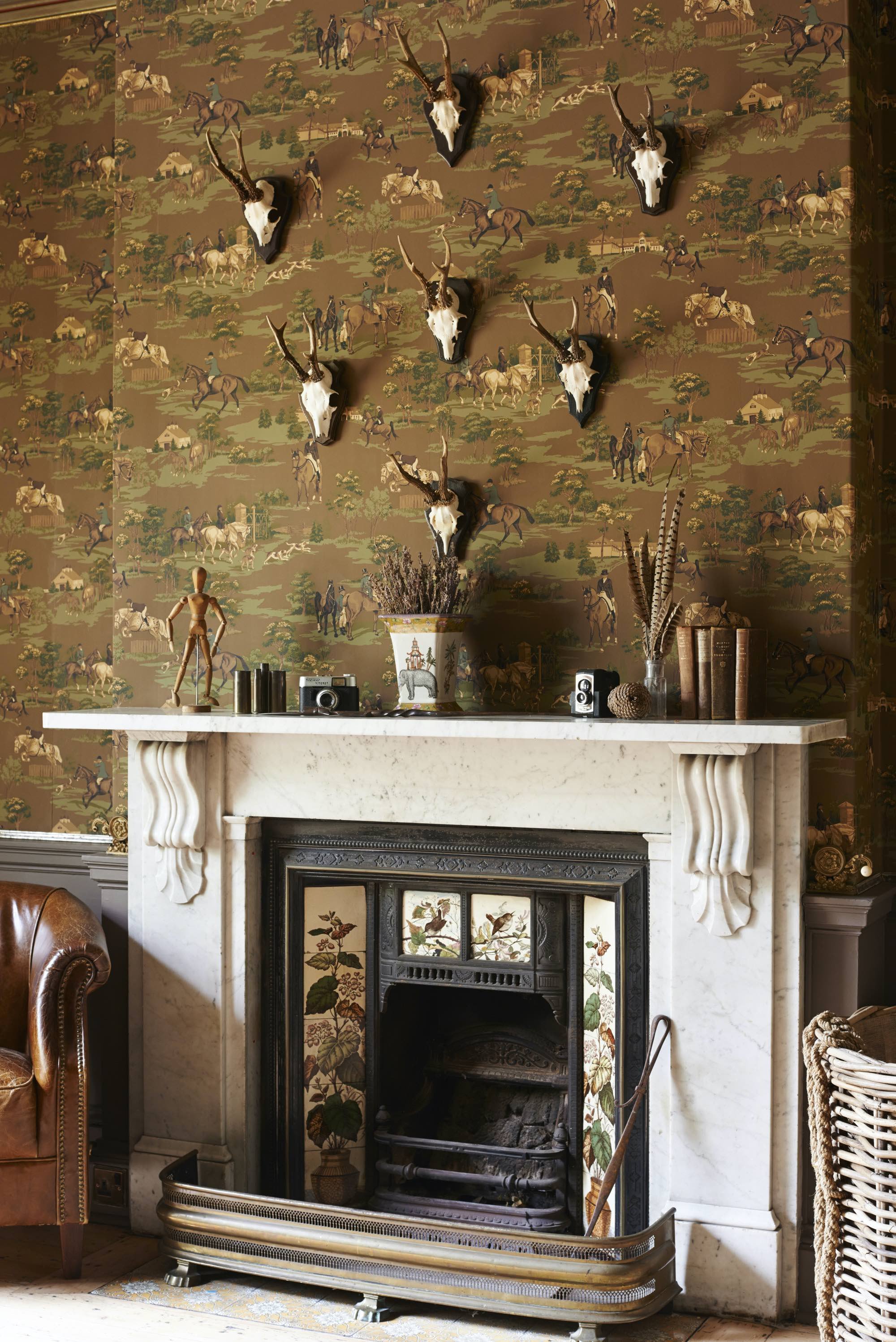 Decorative fireplace with herbs and nature-themed accessories, mounted animal skulls on the wall, and vintage wallpaper depicting a horseback riding scene.