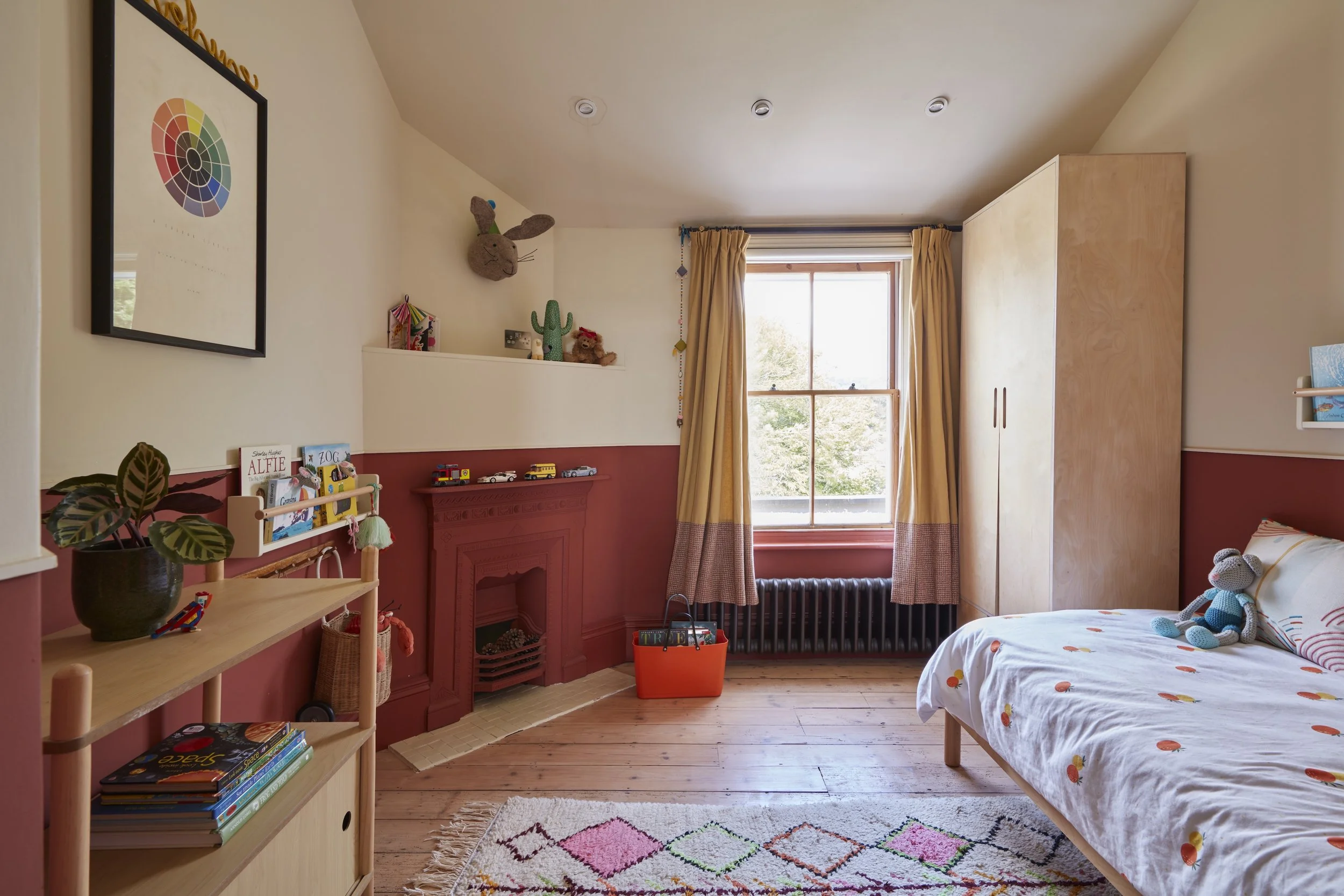 A cozy child's bedroom with a single bed, colorful bedding, and a stuffed toy. Wooden flooring, a window with yellow and pink curtains, and a cream-colored wardrobe. Decor includes small toys, books, a potted plant, artwork, and various plush toys on