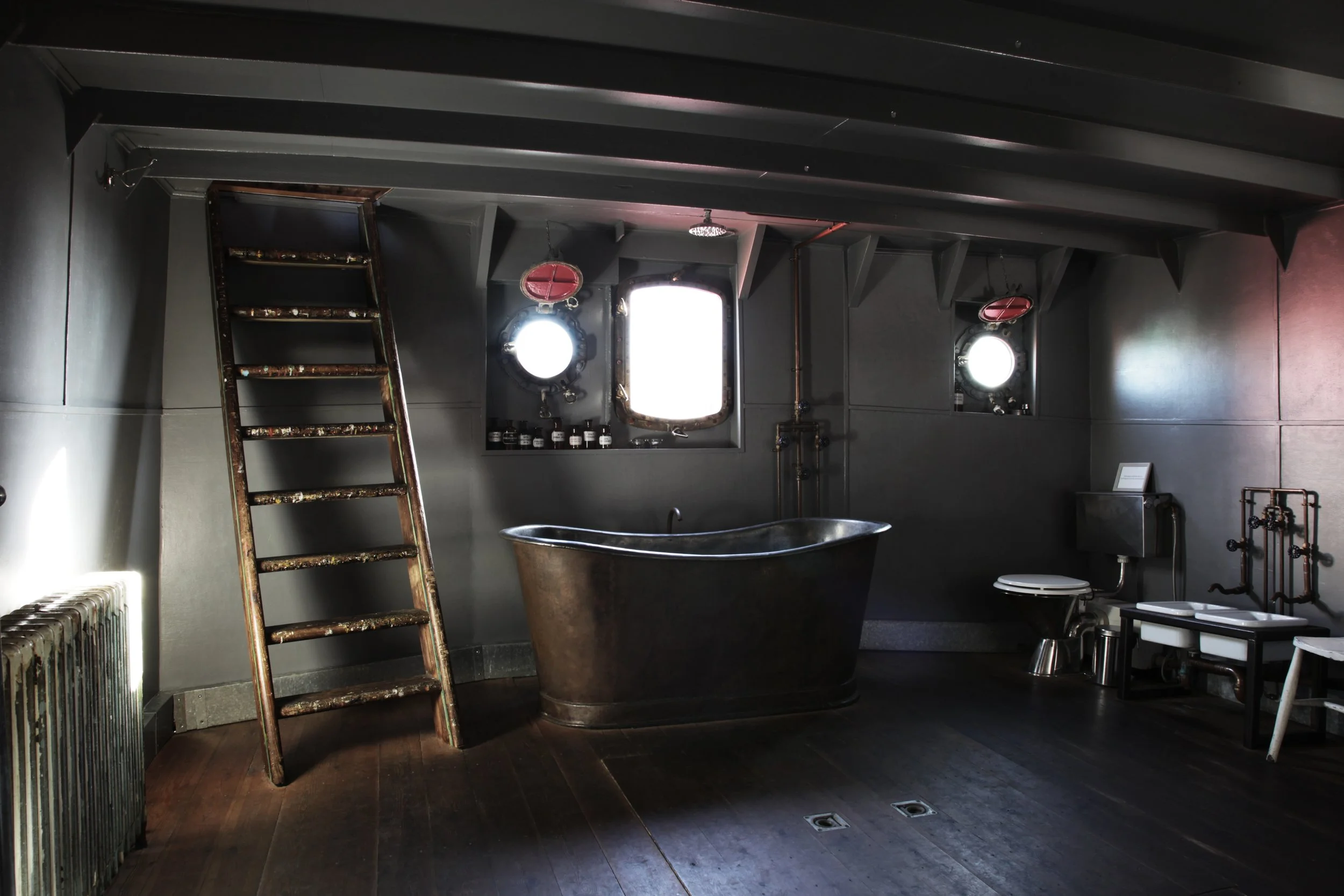 A dark industrial-style bathroom with a vintage clawfoot bathtub, a ladder leaning against the wall, a toilet, and a sink. The room has small round windows with metal fittings and natural light coming through.