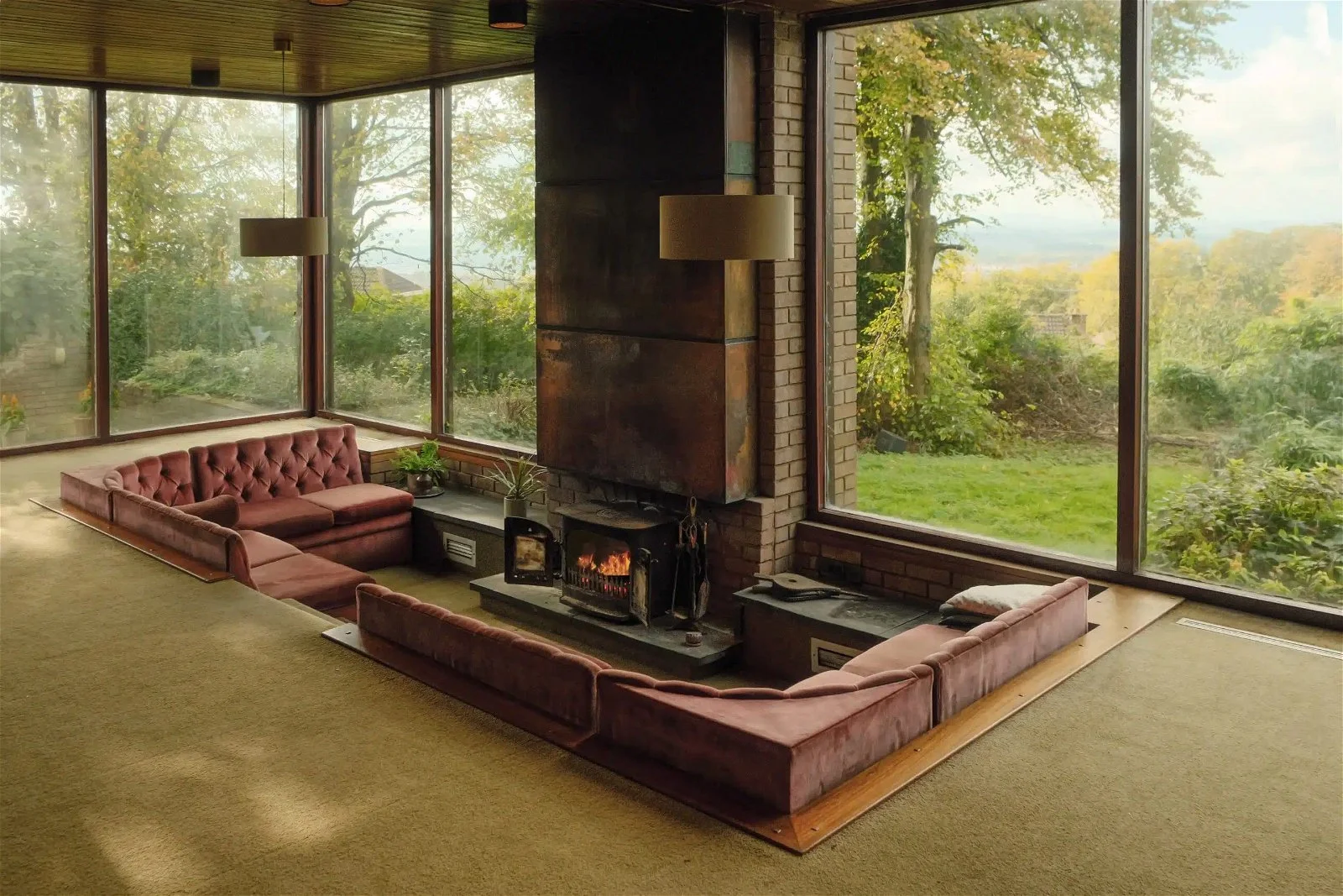 Living room with moss-colored carpet, pink velvet sectional sofa, brick fireplace, large window with views of trees and outdoor landscape, and potted plants inside.