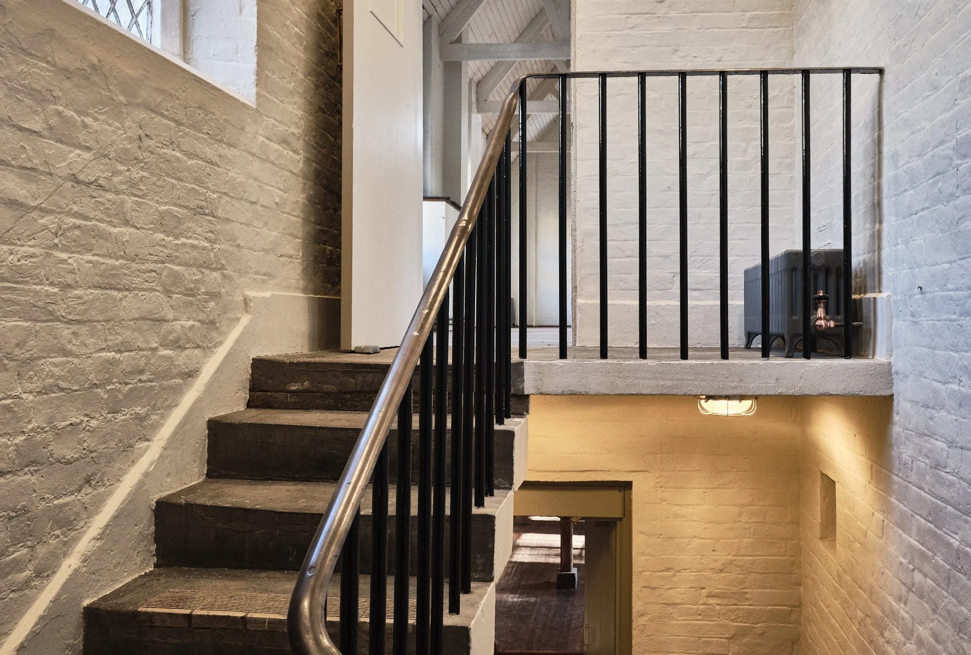 Interior view of a staircase with dark wooden steps, black metal railing, white brick walls, and a small balcony with a black radiator and copper piping.