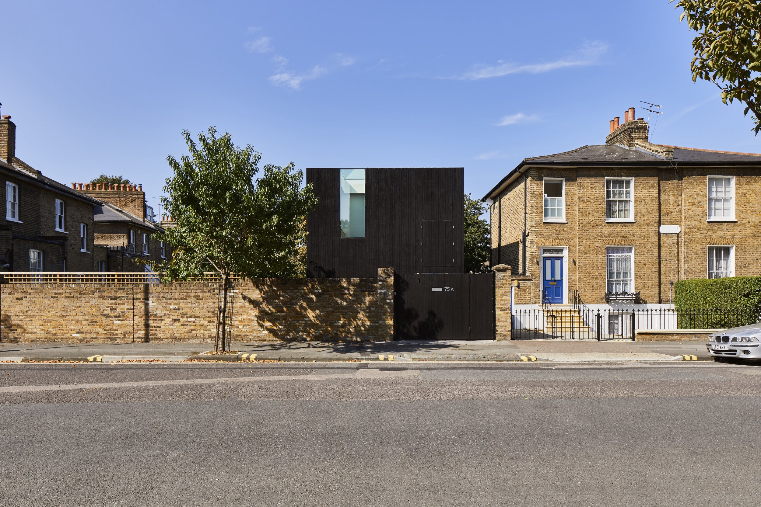 A street view featuring a modern black building with a large window, flanked by traditional brick houses with blue doors and white window frames, under a clear blue sky.