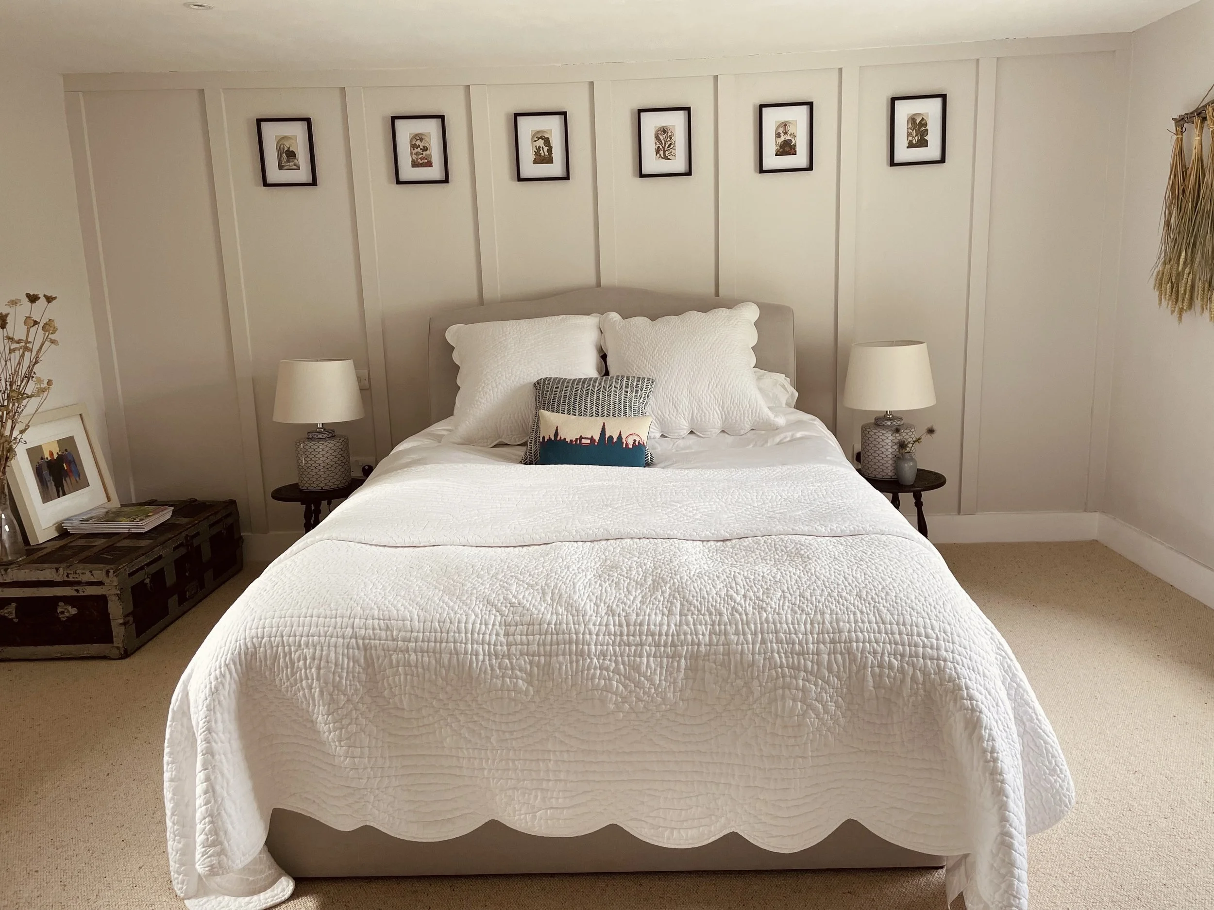 A neatly made bed with white textured bedding, two large white pillows, and a decorative pillow featuring a skyline silhouette with a red sun. There are two white bedside lamps on dark side tables, and framed artwork hanging on the wall behind the be