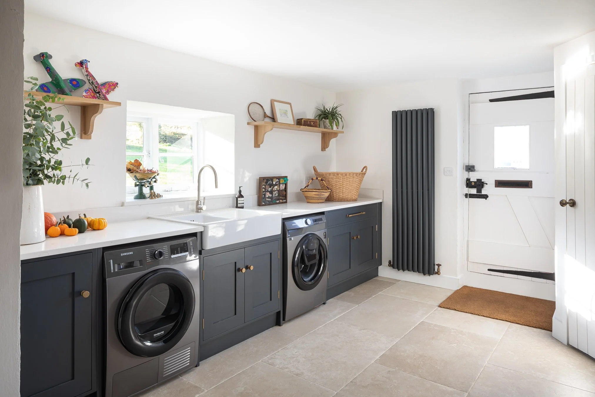 A laundry room with dark blue cabinets, two front-loading washing machines, a white countertop, a white farmhouse sink, and various decorations including pumpkins and shelves with plants and framed artwork.