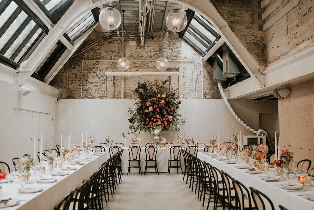 Elegant indoor dining space with long tables set for a meal, floral arrangements, and a large central flower display on a white wall, with pendant lights and skylights.